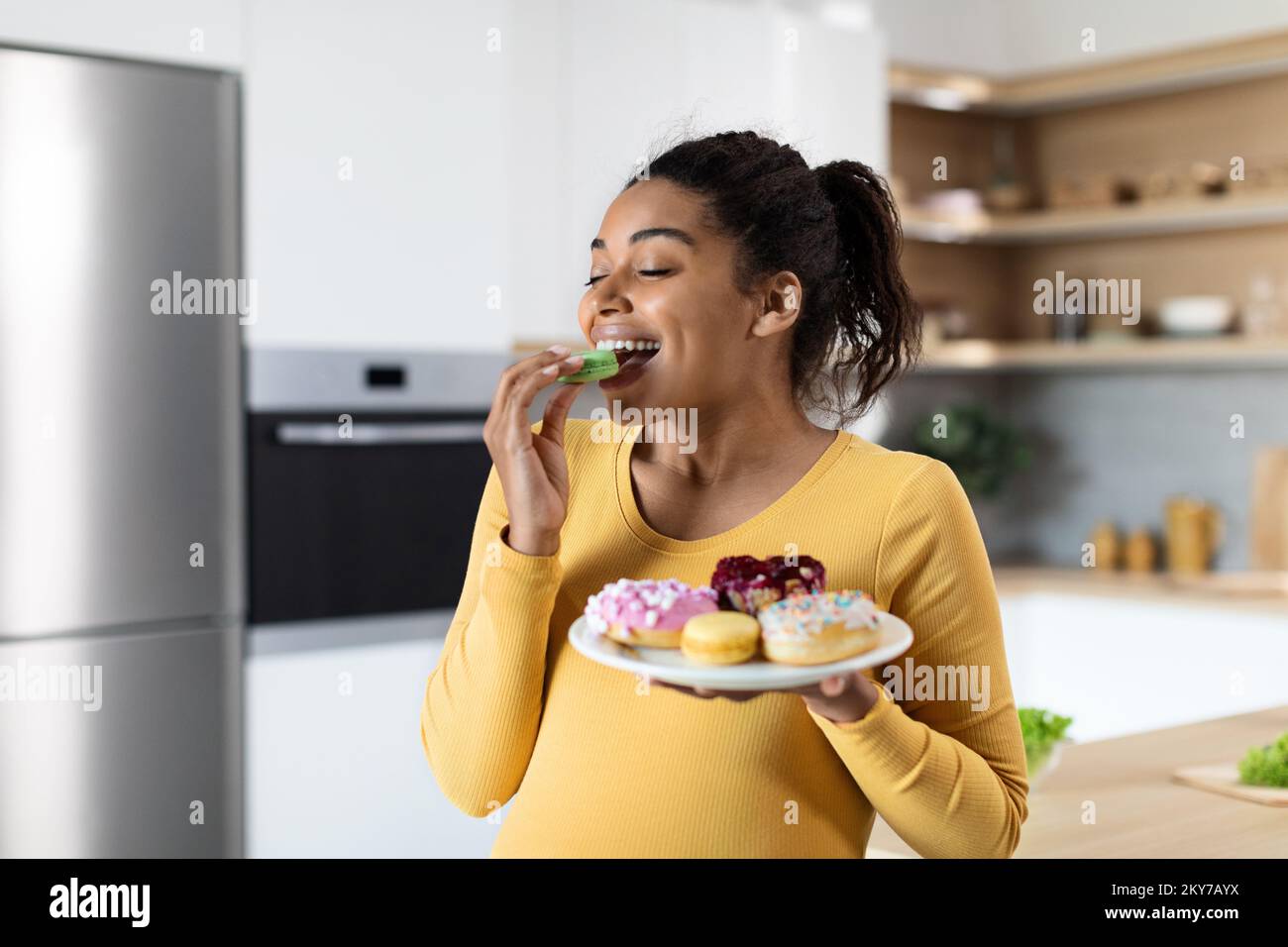 Sorridente affamato giovane donna nera incinta con grande pancia mangia dolci, tenere piatto con biscotti Foto Stock