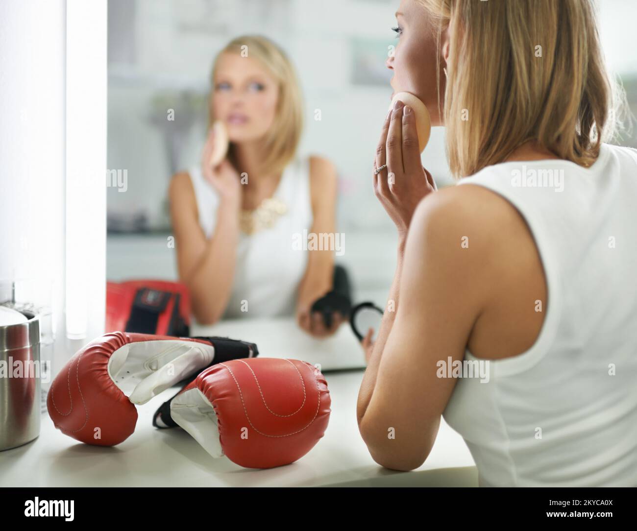 Abbastanza duro. Una giovane donna alla moda che applica il make-up con un paio di guanti di boxe che si trovano accanto a lei. Foto Stock