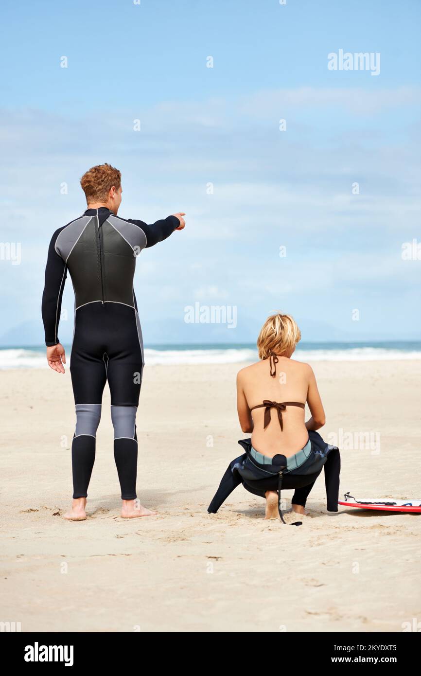 In attesa dell'onda perfetta. Due surfisti seduti sulla spiaggia mentre uno punta verso l'oceano. Foto Stock