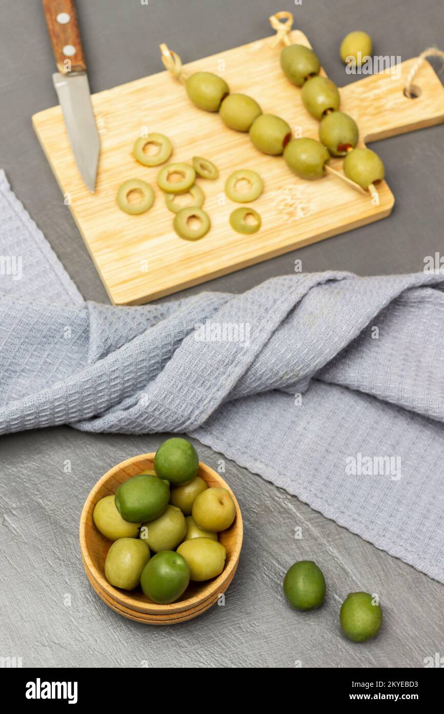 Olive verdi in una ciotola di legno. Tovagliolo grigio. Coltello e olive tritate su un tagliere. Giacitura piatta. Sfondo grigio. Foto Stock
