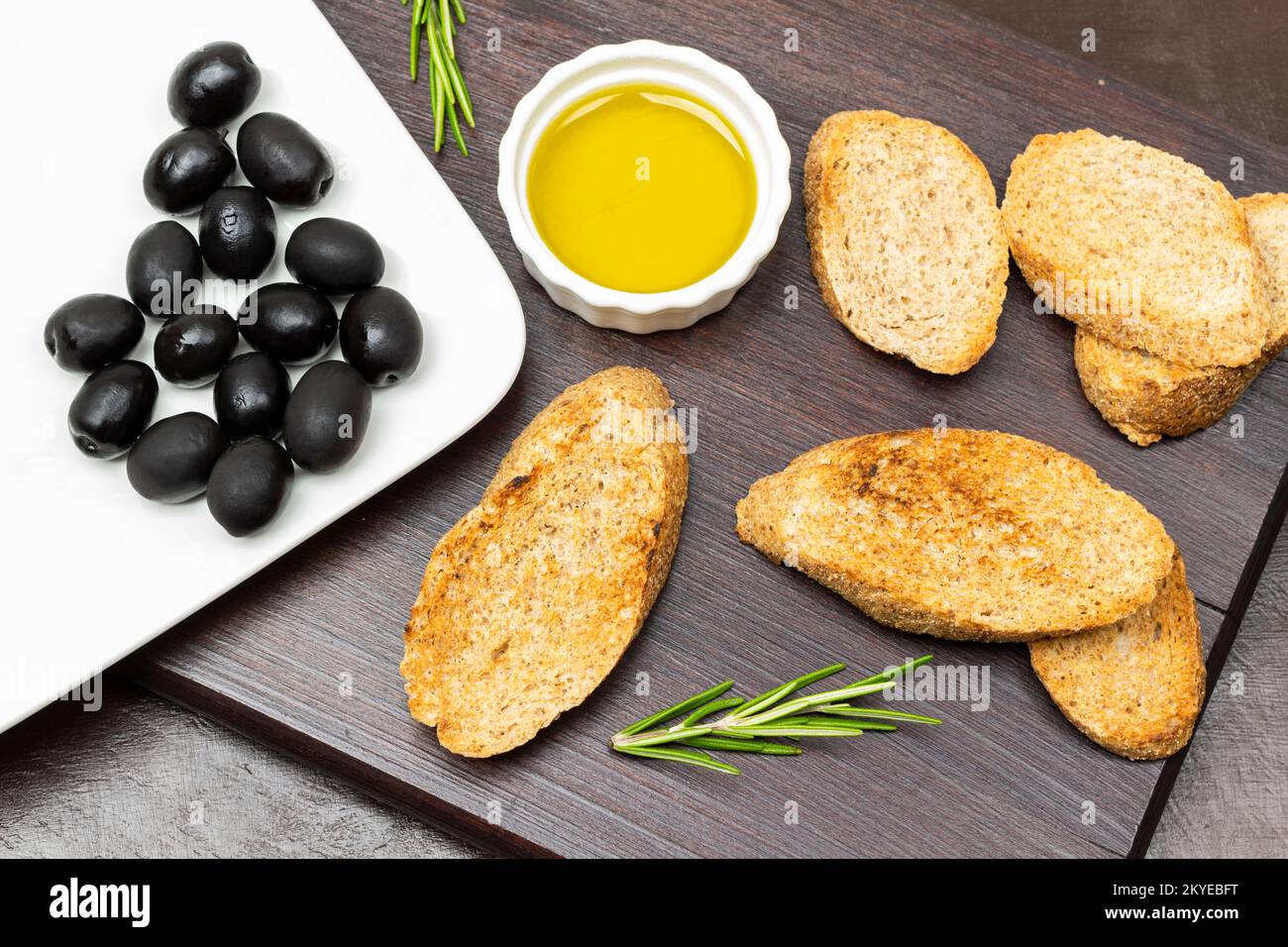 Olive nere su un piatto bianco. Tostare e olio d'oliva in un recipiente. Giacitura piatta. Sfondo marrone. Foto Stock