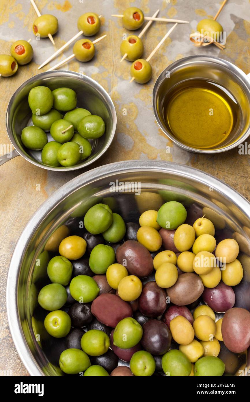 Diverse varietà di olive in una ciotola di metallo. Olive verdi e olio in ciotole. Olive sugli spiedini. Giacitura piatta. Fondo in metallo arrugginito. Foto Stock