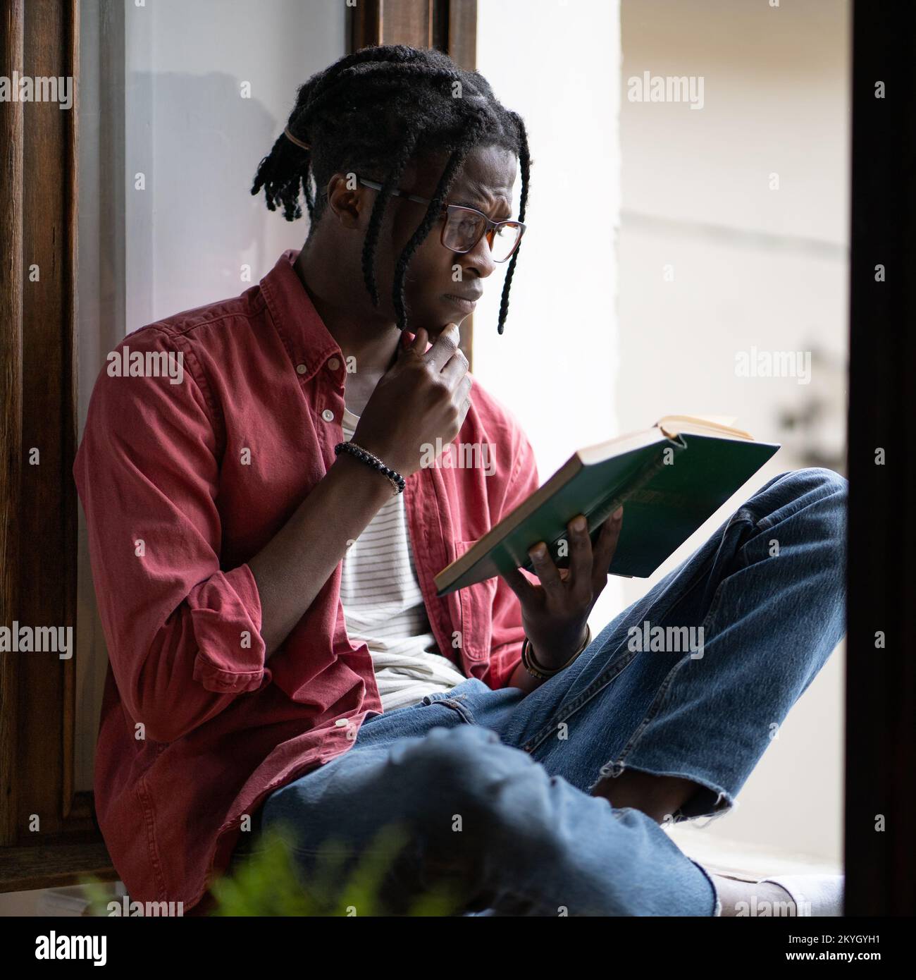 Concentrato inquisitivo afro americano studente libro di lettura si siede sul windowsill vicino alla finestra Foto Stock