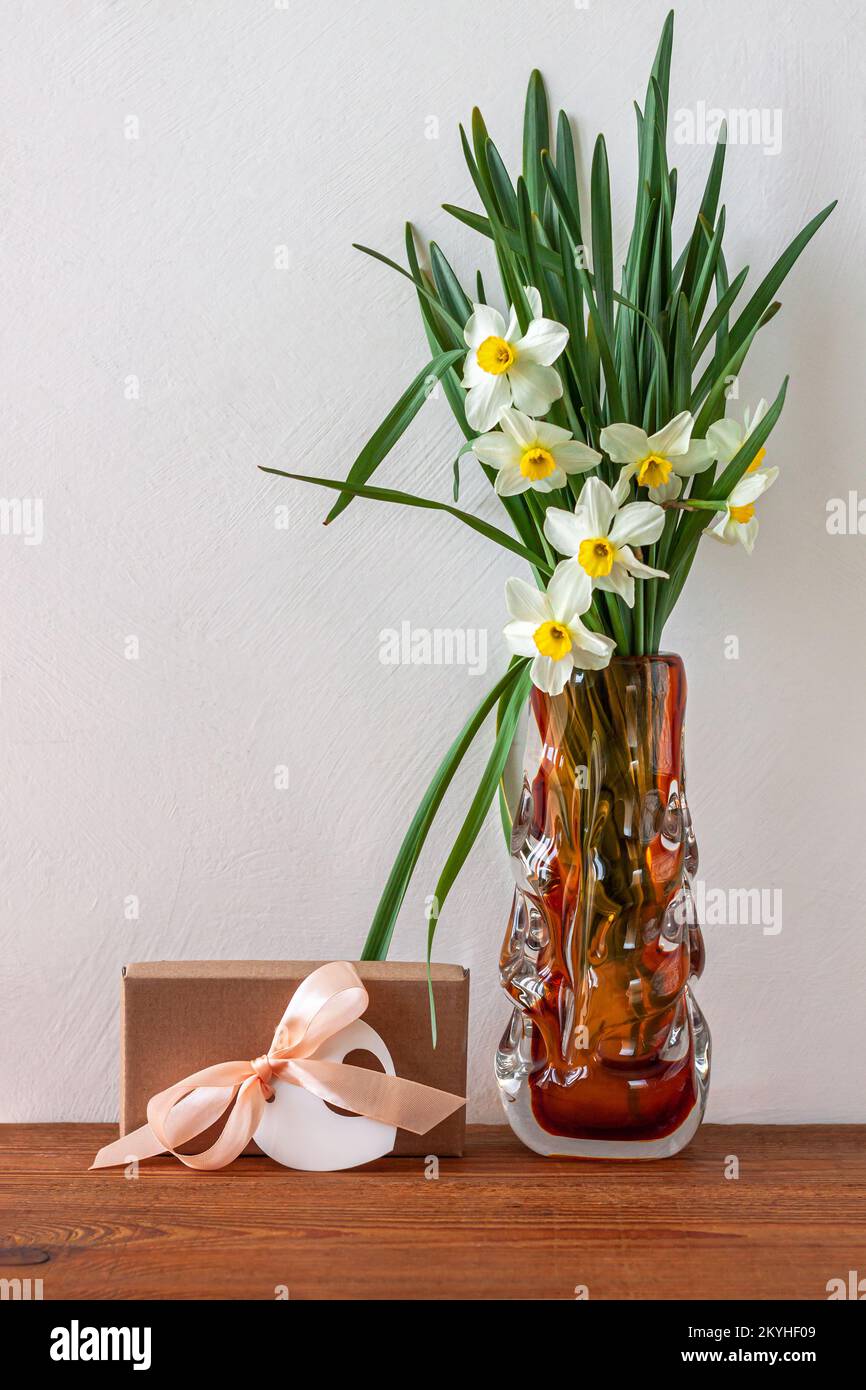 Fiori di narcisi bianchi e scatola regalo marrone, biglietto d'auguri con nastro rosa su sfondo chiaro. Luogo per il testo 8 marzo buon giorno delle madri delle donne. Bouqu Foto Stock