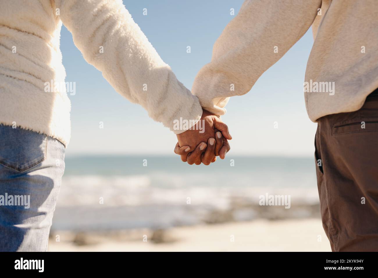Vista posteriore di una coppia matura che tiene le mani in piedi su un ponte di legno sulla spiaggia. Romantica coppia senior che fa una rinfrescante vacanza al mare Foto Stock