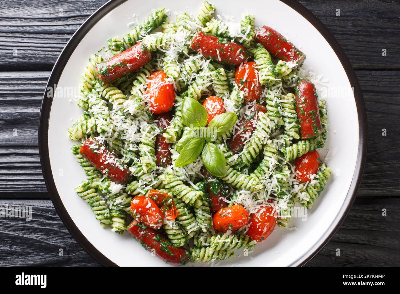 Pranzo abbondante Fusilli pasta con salsicce, pomodori, salsa di pesto verde e formaggio primo piano in un piatto sul tavolo. Vista orizzontale dall'alto Foto Stock
