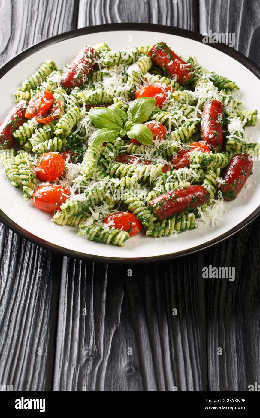 Fusilli con salsicce, pomodori, pesto e parmigiano in un piatto sul tavolo. Verticale Foto Stock