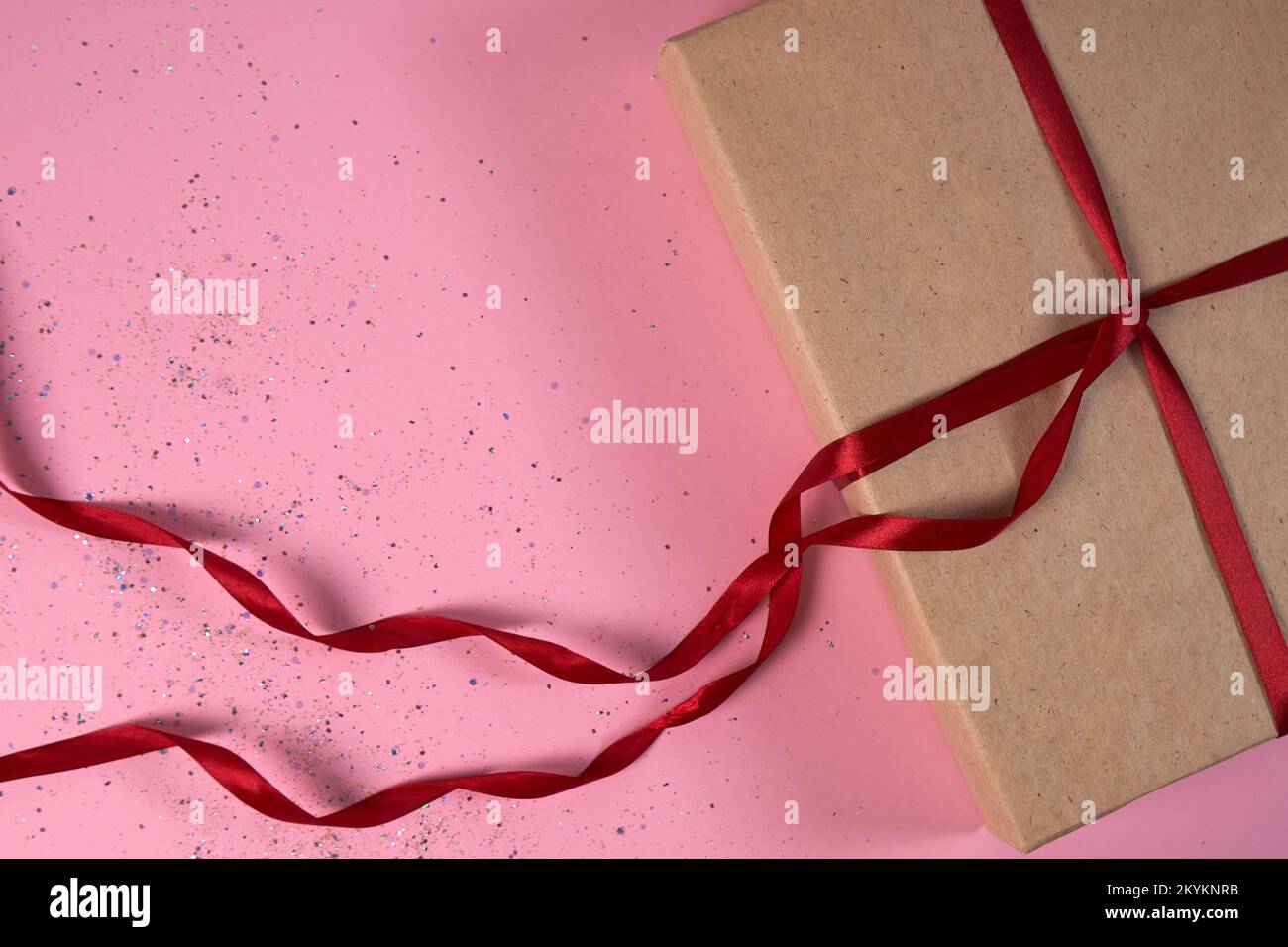 Confezione regalo in carta kraft con nastri di raso rosso su sfondo rosa con paillettes. Il concetto di Capodanno, Natale, Vacanze, San Valentino, matrimonio. Foto Stock