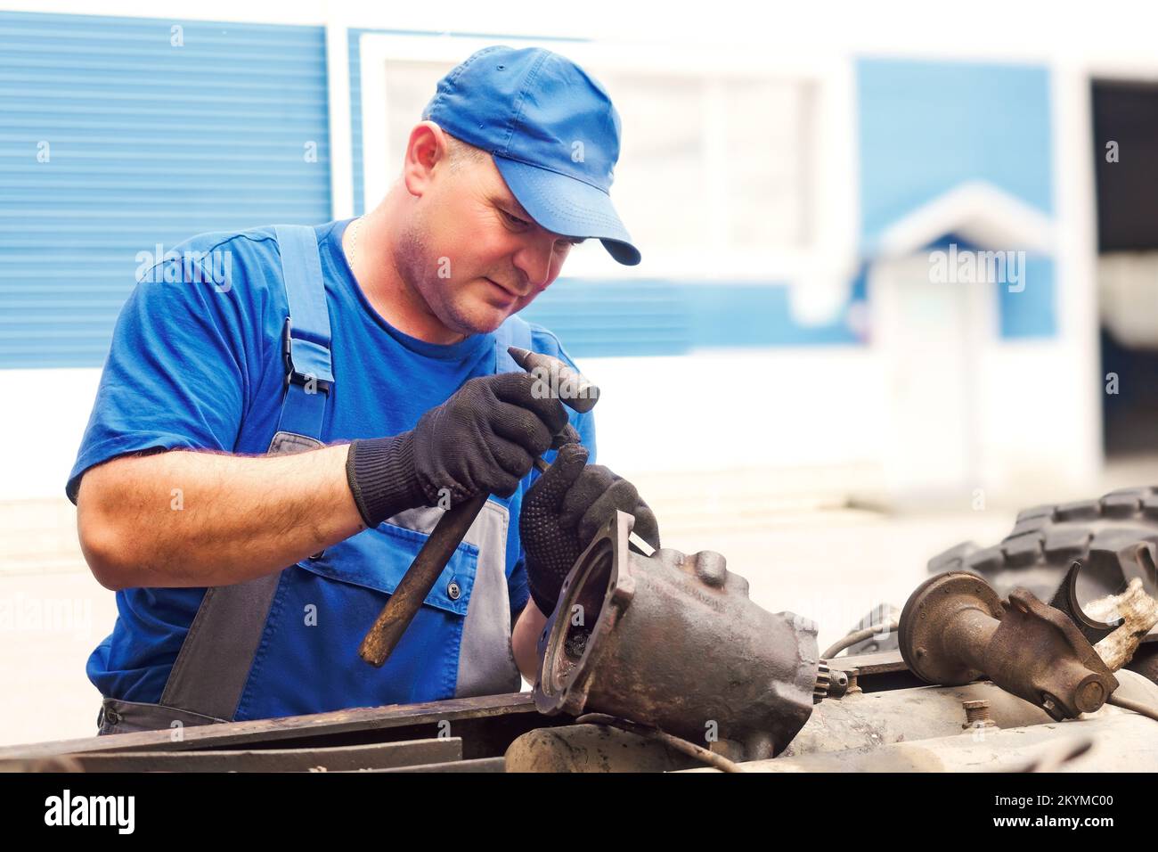 Meccanico in tute e cappellino da baseball ripara trattore o camion il giorno d'estate. Vista frontale. Assistenza professionale nella manutenzione di attrezzature pesanti. Flusso di lavoro autentico. Foto Stock