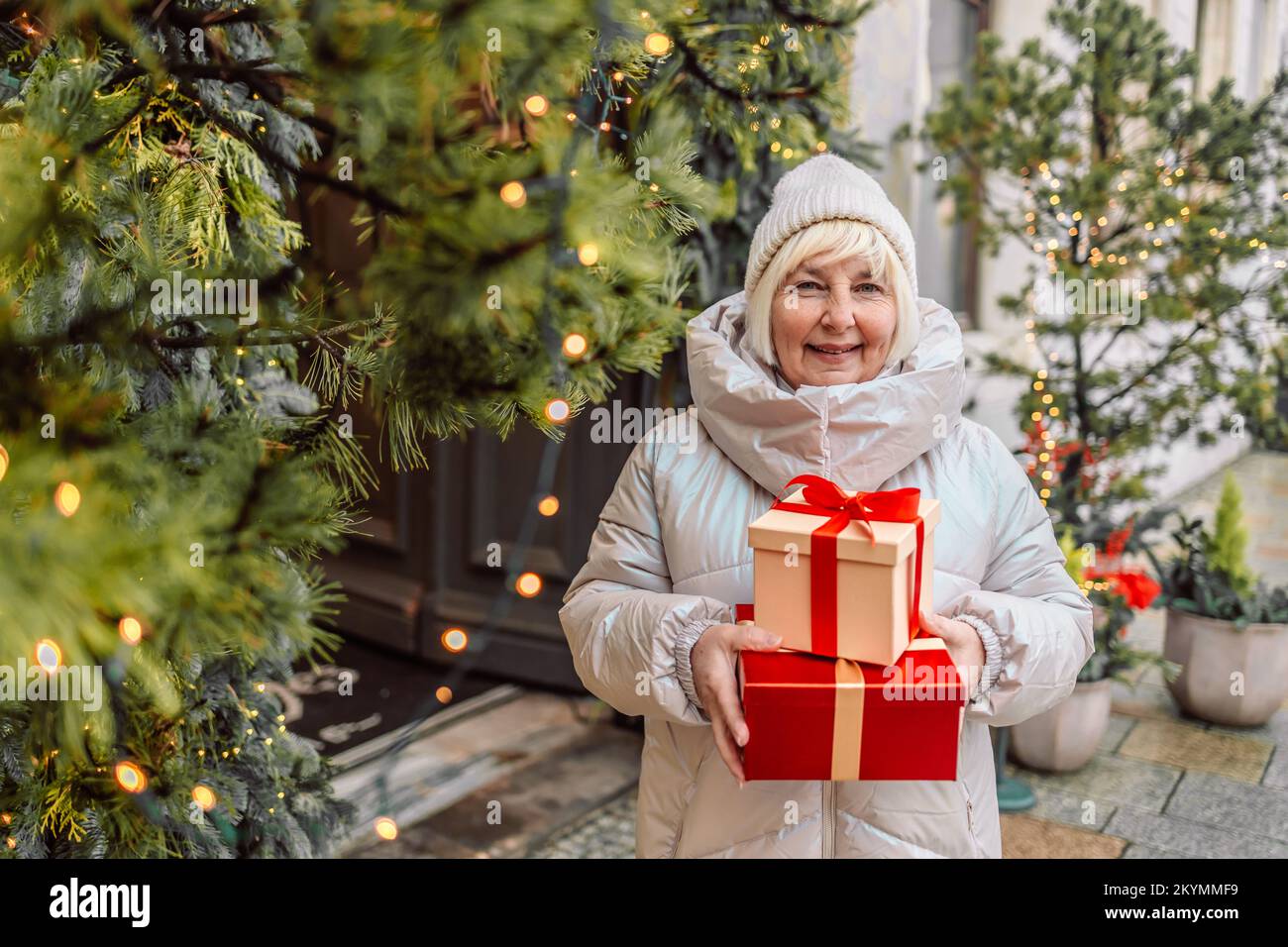 Buon Natale e buone feste. Donna anziana che si diverte con una scatola regalo a sorpresa presente vicino all'albero di Natale all'aperto al mercatino di Natale in città Foto Stock