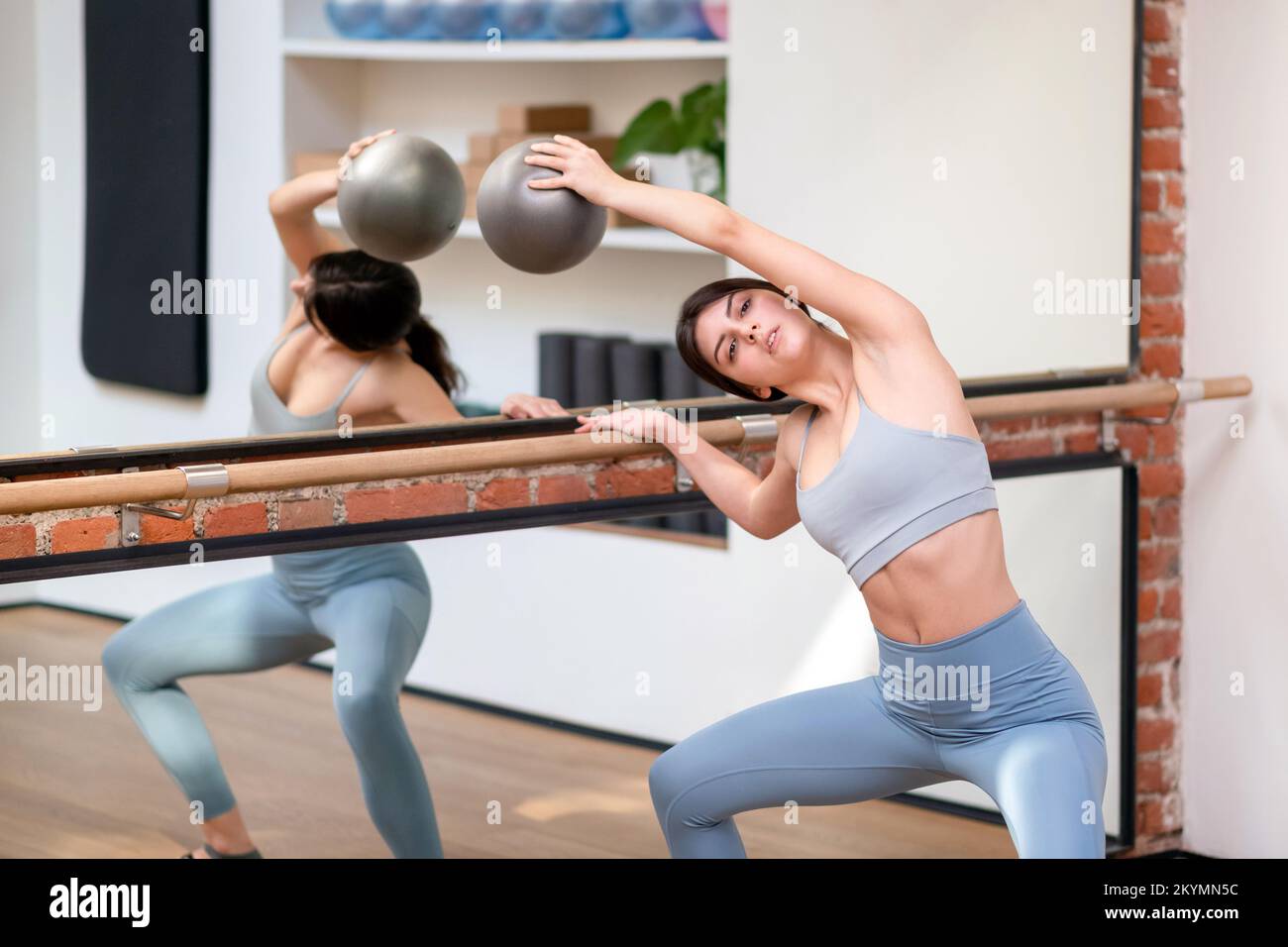 Sottile giovane brunetta con palla che si piega da parte e facendo squat plie vicino corrimano e specchio durante la prova di balletto in studio Foto Stock