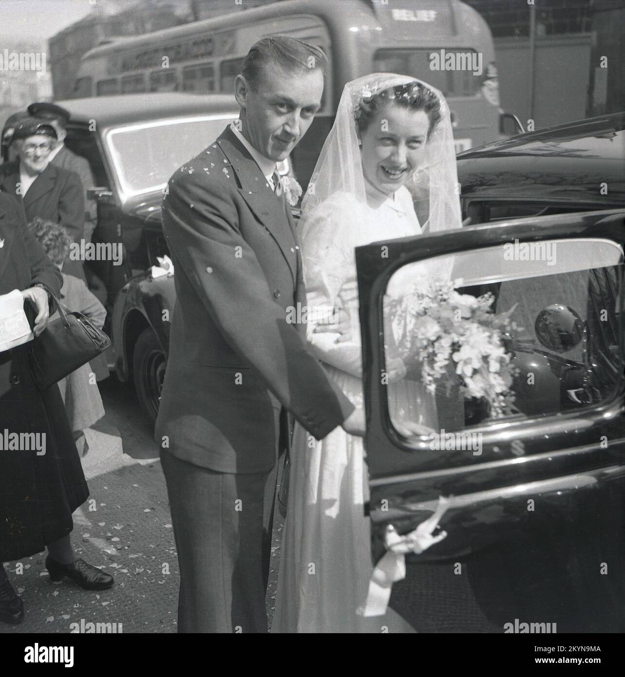 1953, storico, fuori su una strada trafficata di Londra, una coppia appena sposata stare insieme per una foto, in procinto di entrare nella porta aperta di una macchina in attesa per andare al ricevimento di nozze, Inghilterra, Regno Unito. Foto Stock