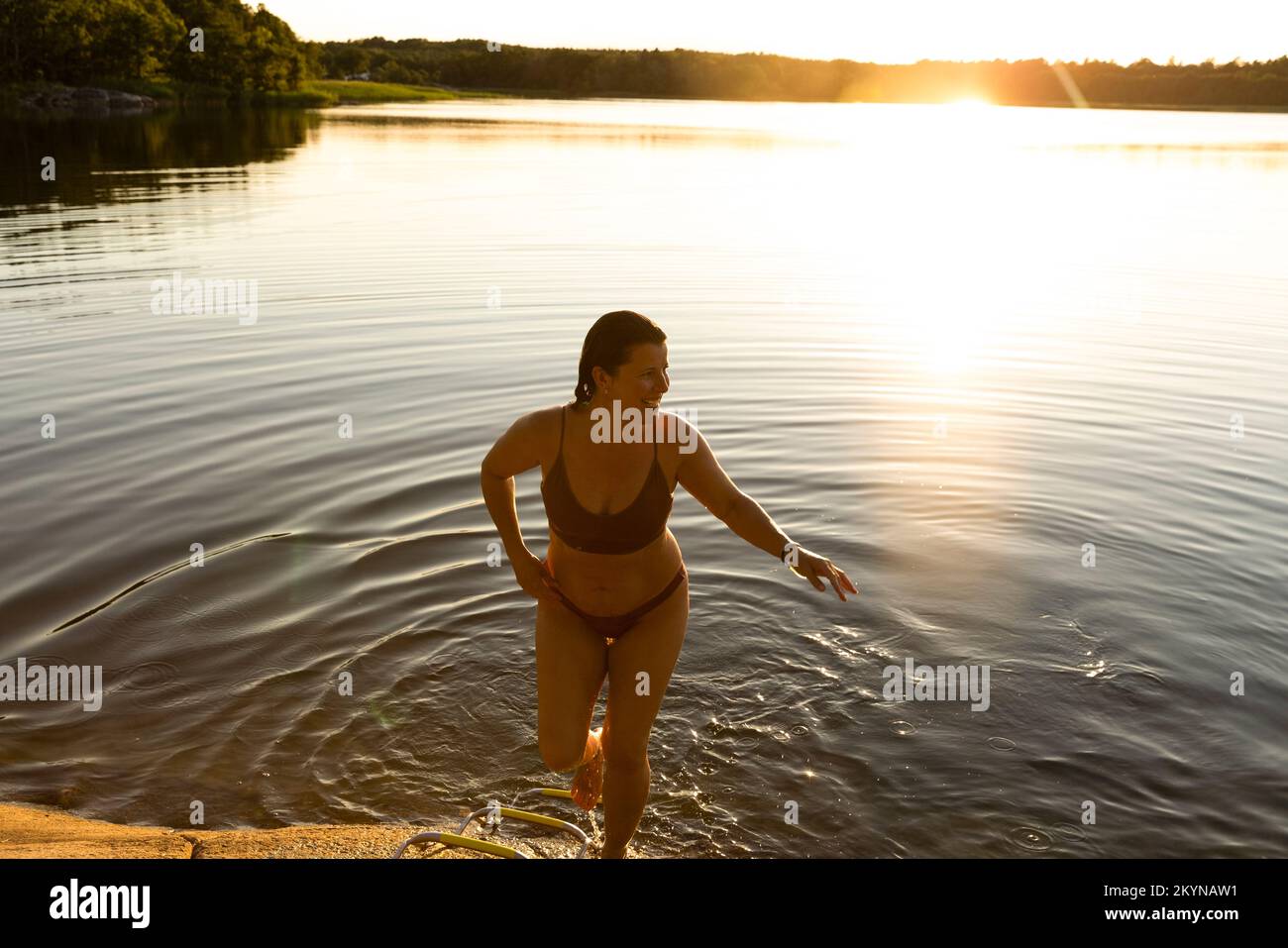 Donna felice in bikini che cammina fuori dal lago dopo aver nuotato durante il tramonto Foto Stock