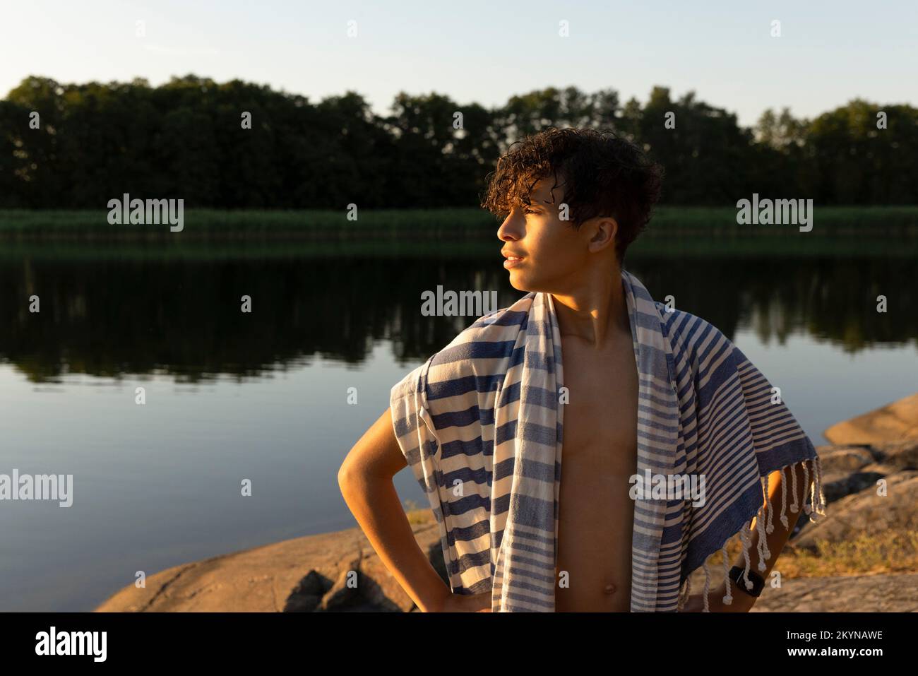 Ragazzo adolescente con asciugamano che si affaccia sul lago durante il tramonto Foto Stock