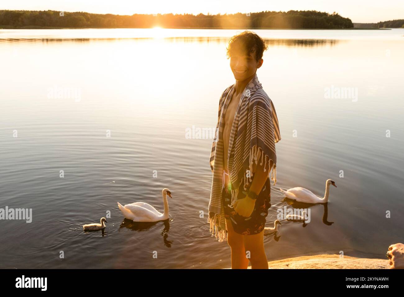 Ritratto di ragazzo felice in piedi vicino al lago con cigni che nuotano sullo sfondo Foto Stock