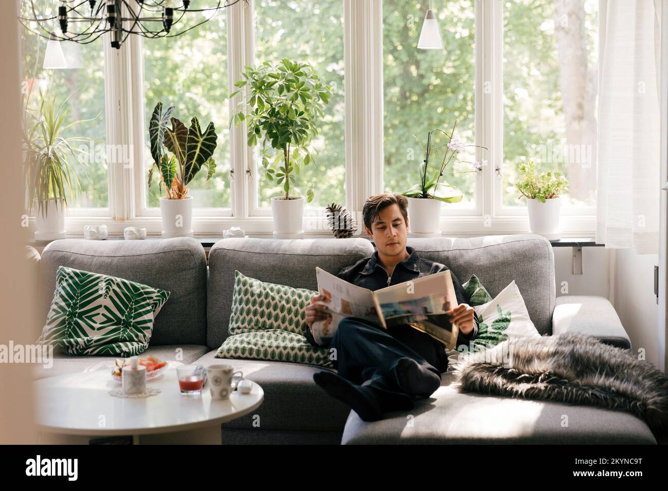 Giovane uomo che legge il giornale mentre si siede sul divano nel soggiorno di casa Foto Stock