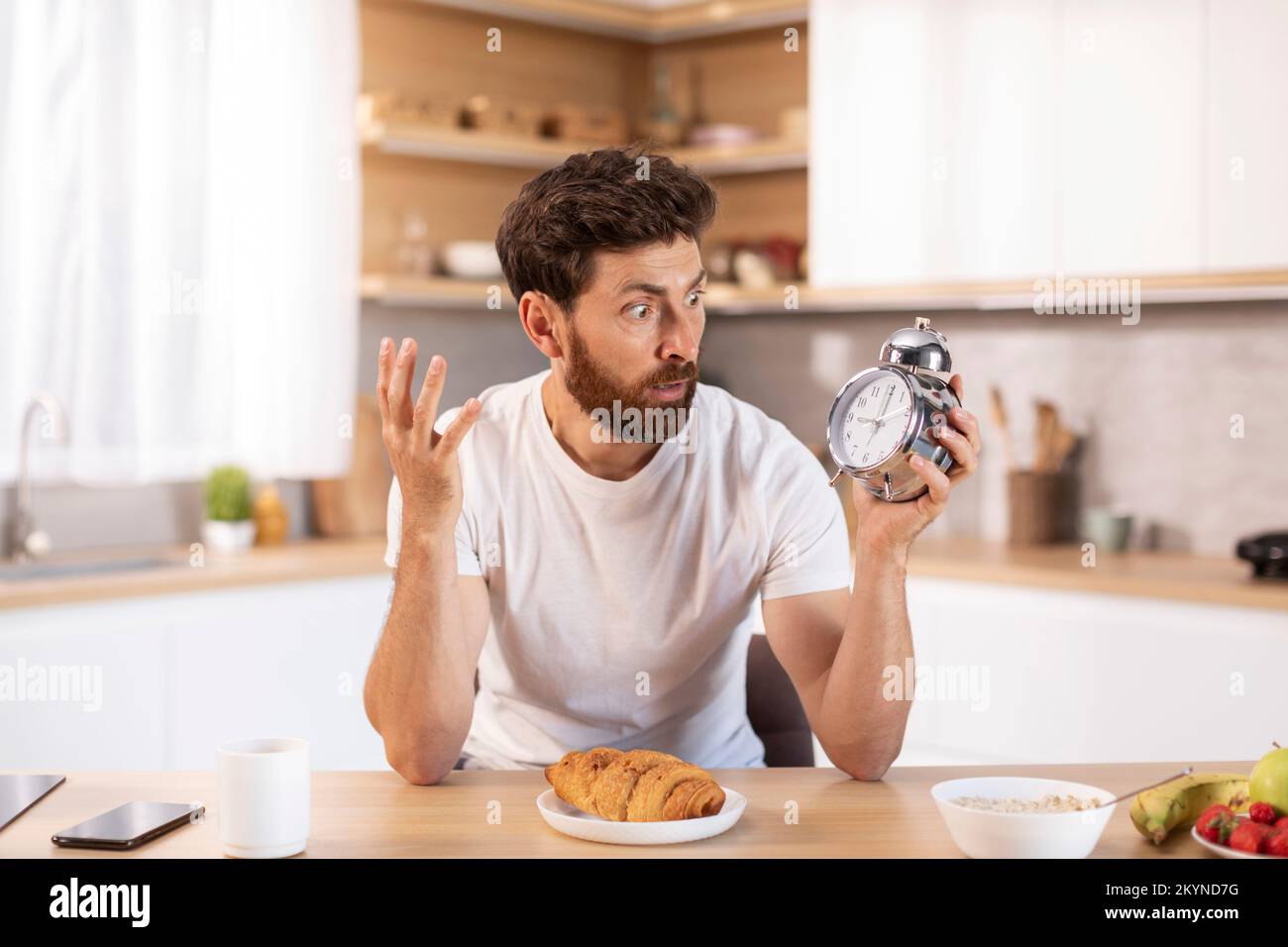 Scioccato triste uomo caucasico adulto con barba in t-shirt bianca guarda la sveglia e tardi per incontrarsi o lavorare Foto Stock