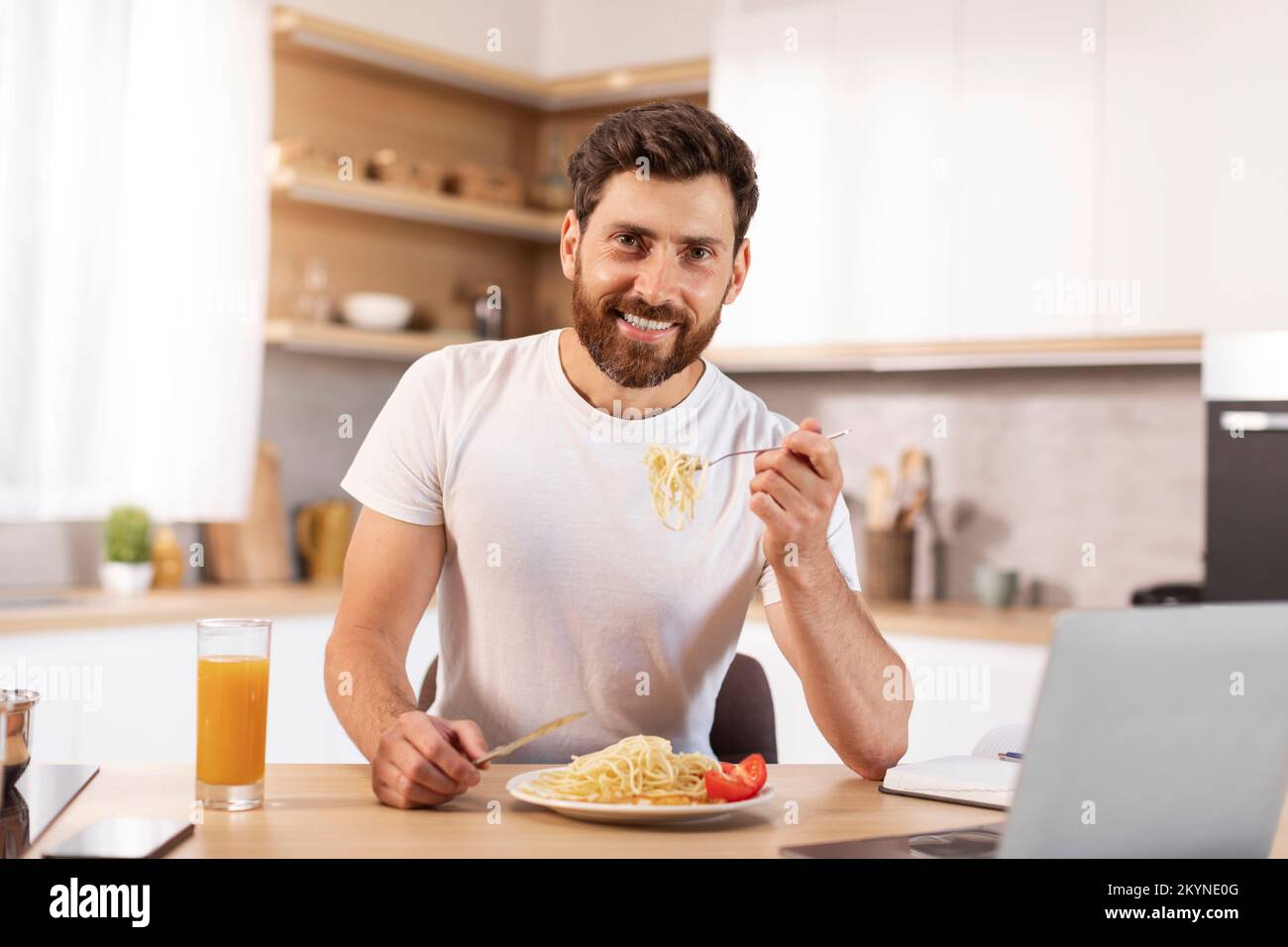 Buon buon uomo caucasico maturo sopportato mangiare pasta in interni cucina minimalista con computer portatile Foto Stock