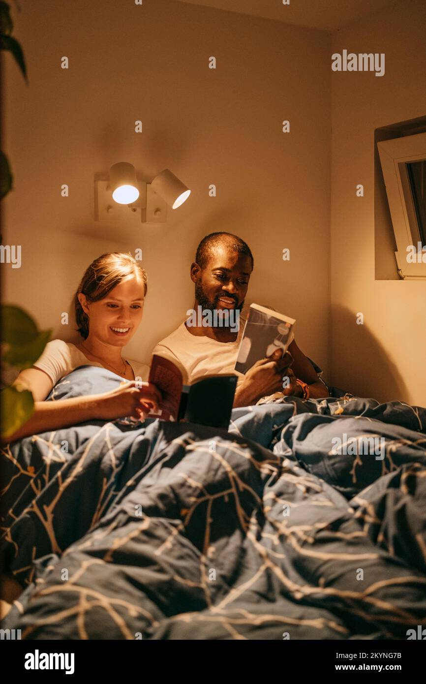 Uomo e donna che leggono libri mentre riposano sul letto con coperta a casa Foto Stock