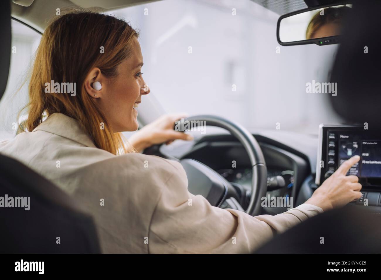 Donna d'affari sorridente che indossa cuffie auricolari wireless utilizzando il pannello di controllo del cruscotto in auto Foto Stock