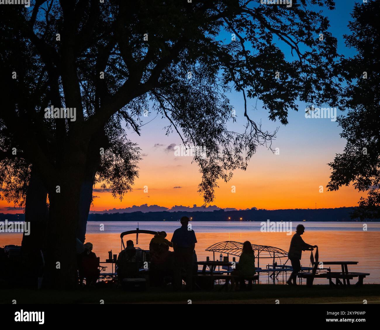 Silhouette di persone e alberi e barche a riva del lago con un tramonto arancione in lontananza al crepuscolo. Foto Stock