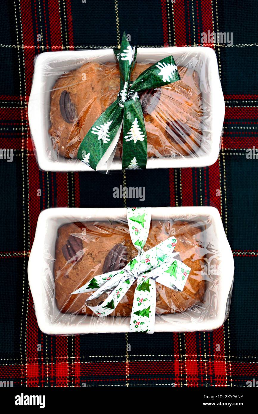 Mini pagnotte di banana appena sfornata con noci pecan avvolti in un involucro di plastica trasparente con fiocchi festosi su sfondo tartan in formato verticale. Foto Stock