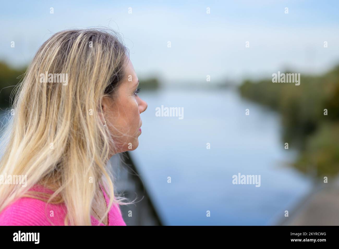Donna penosa di mezza età che indossa un maglione rosa e rossetto rosa guarda sopra la spalla a un fiume o lago in autunno Foto Stock