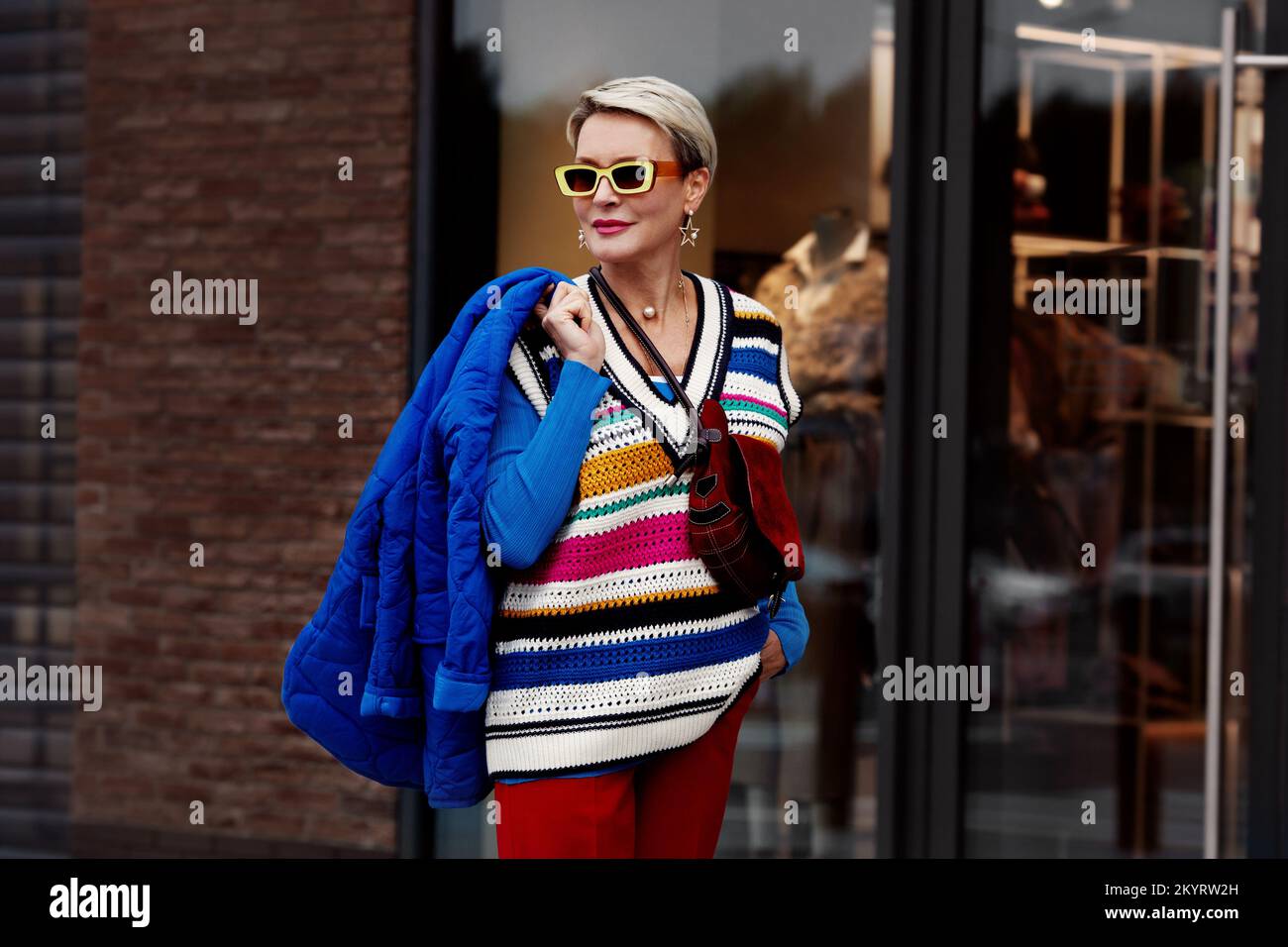 Moda Street stile autunno o primavera. La modella indossa giacca e maglione blu, doratura a righe, pantaloni rossi e occhiali da sole gialli. Donna in posa all'esterno Foto Stock