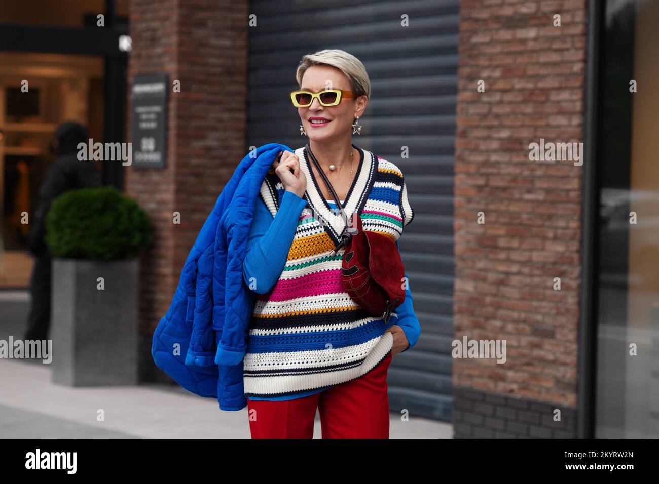Sorridente Happy Model indossa giacca e maglione blu, doratura a righe, pantaloni rossi e occhiali da sole gialli. Moda Street stile autunno o primavera. Donna p Foto Stock