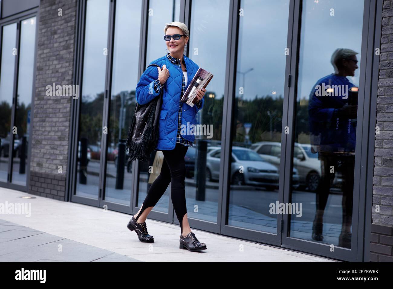 Donna alla moda con i capelli corti che camminano strada vestita in elegante vestito casual, giacca e occhiali da sole colorati blu, borsa e stivale in pelle nera Foto Stock