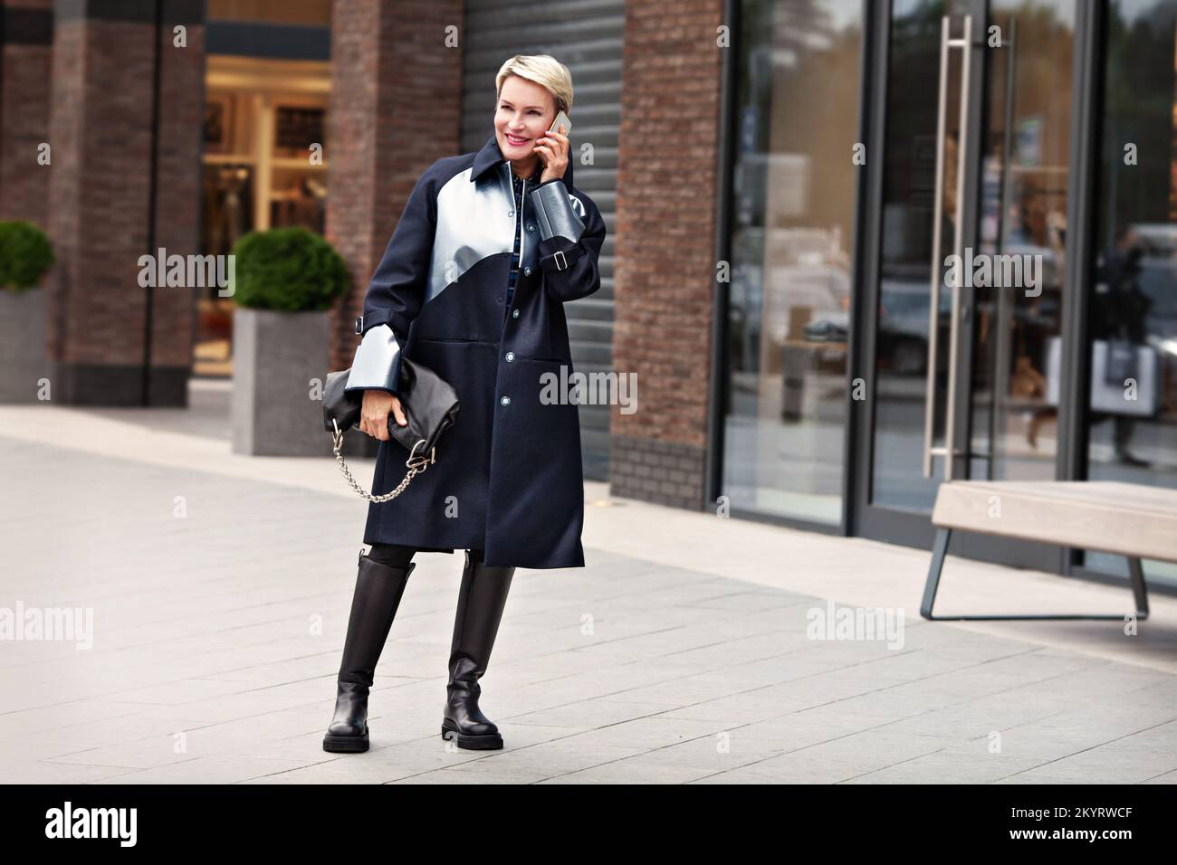 Moda Donna in abito alla moda, cappotto nero, stivaletti in pelle, leggings. Elegante modello alla moda è parlare telefono e sorridente sulla strada cittadina vicino Foto Stock