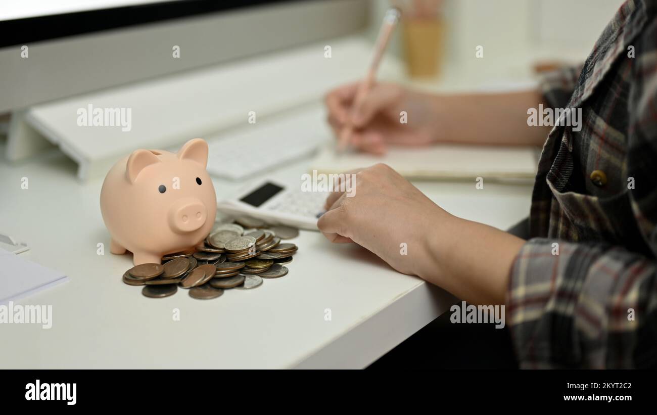 Immagine ravvicinata, Piggy bank e monete sul tavolo con la donna che gestisce e fa i conti mensili di entrate e spese. Foto Stock