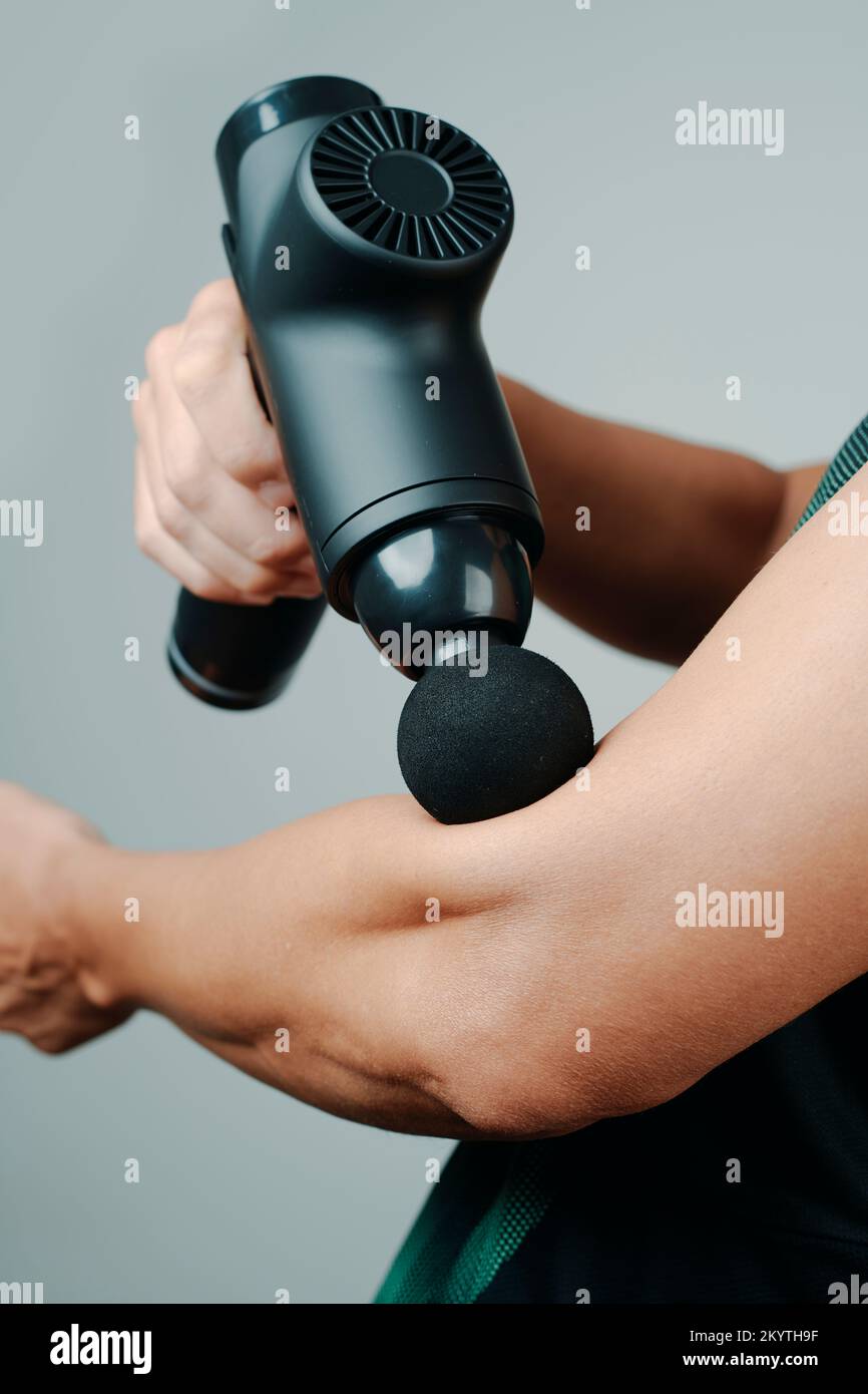 primo piano di un giovane, in abbigliamento sportivo, con una pistola da massaggio per massaggiare i muscoli del braccio, vicino al gomito Foto Stock