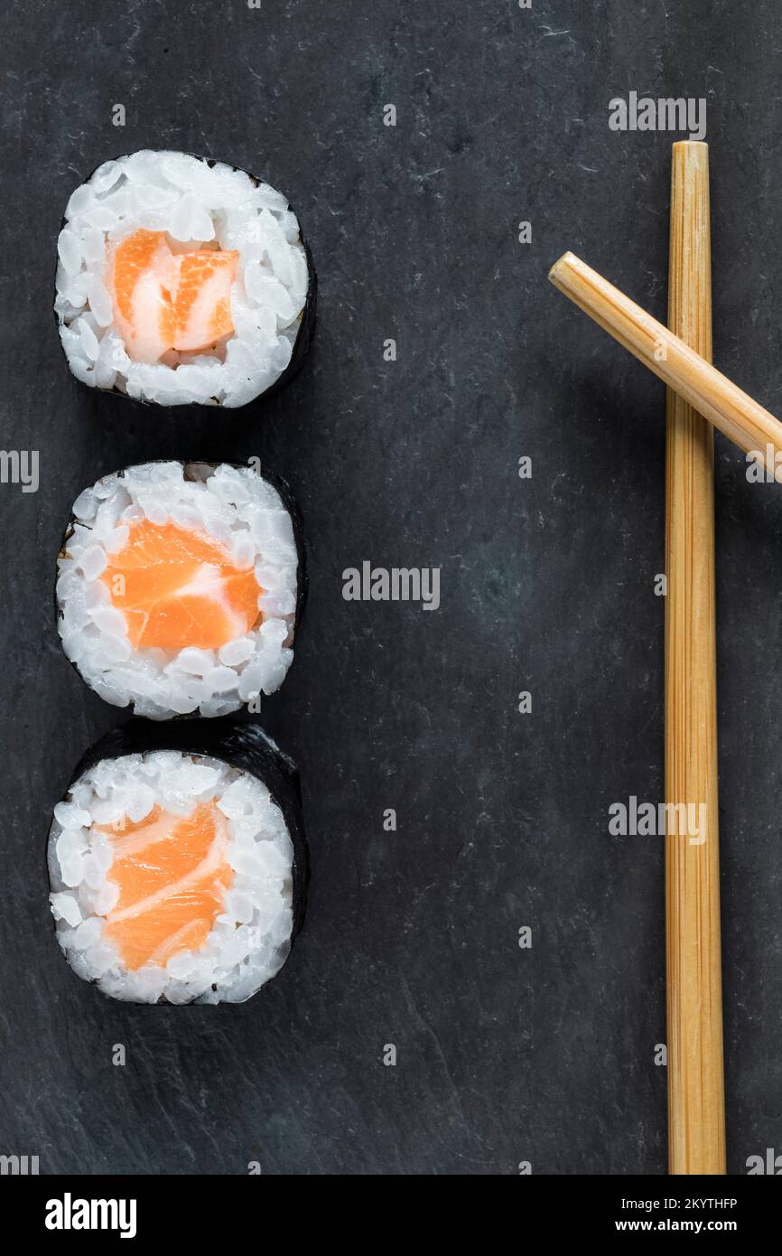 Vista dall'alto dei makis di salmone allineati su sfondo di pietra nera con bacchette accanto ad esso. Orientale, cucina giapponese. Da asporto. Ripresa con obiettivo macro. Foto Stock