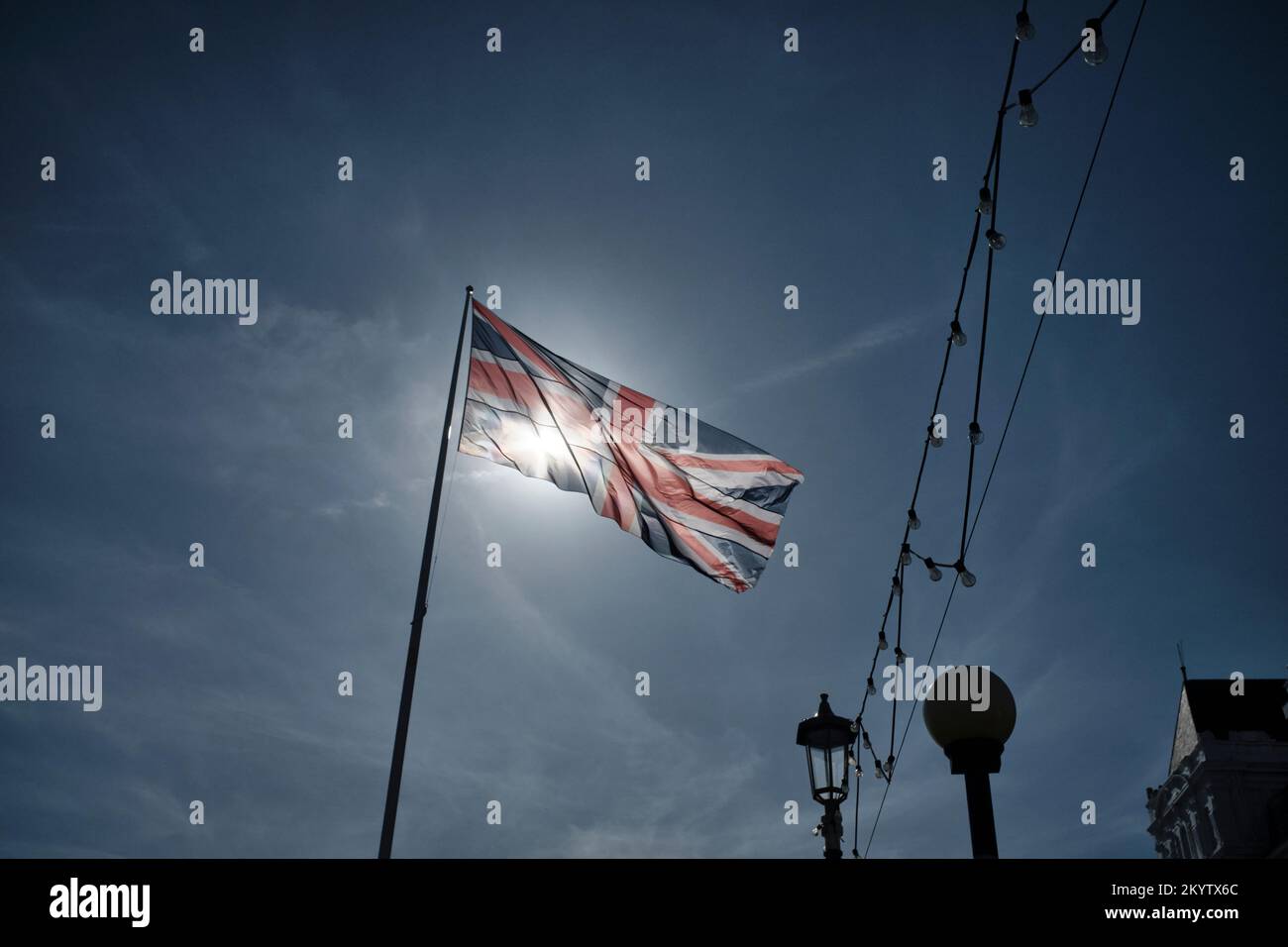 Bandiera di Union Jack retroilluminata dal sole su un flagpole rivolto a destra vicino a una serie di luci festone all'aperto Foto Stock