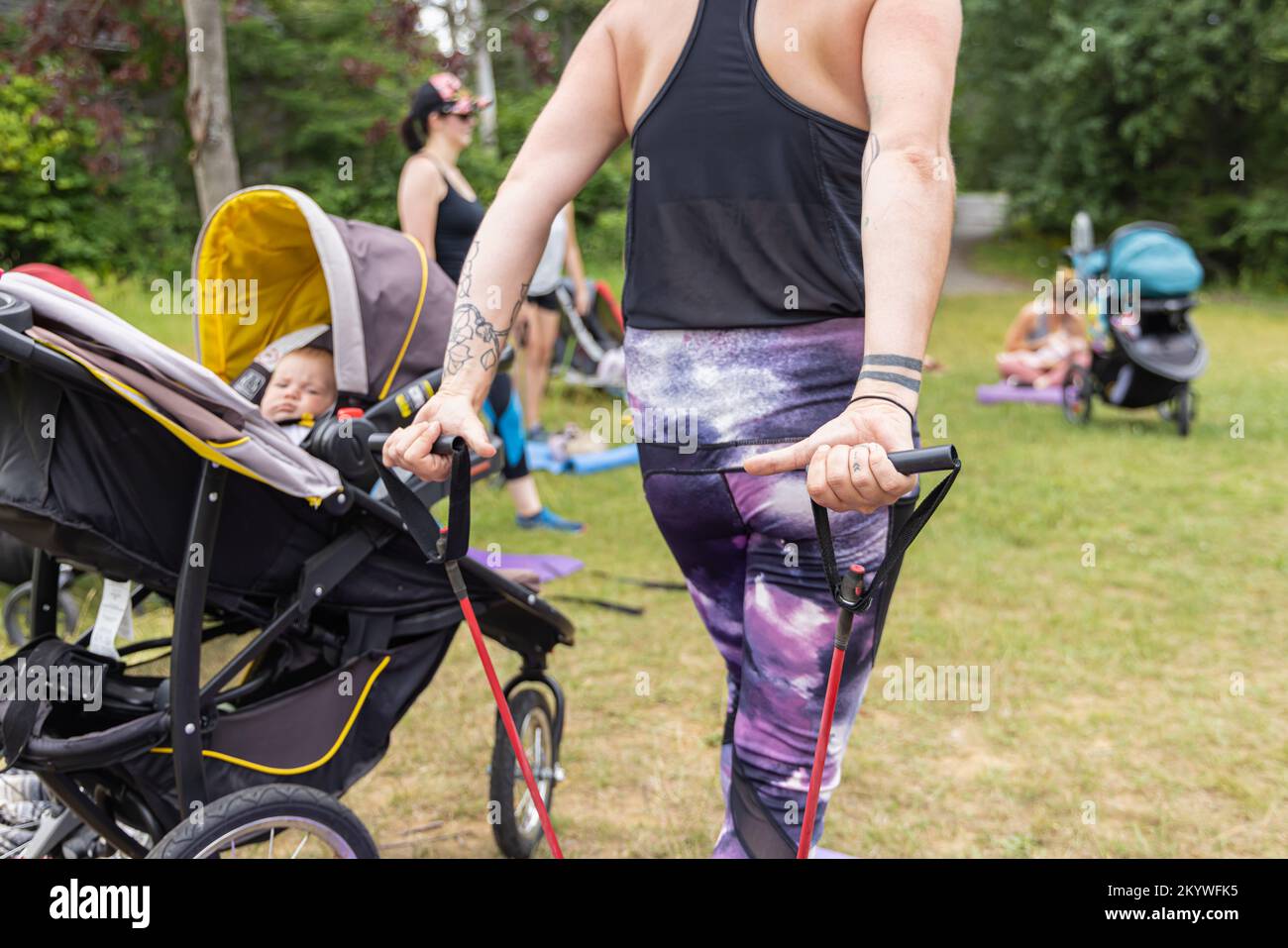 Foto sul retro scattata da una donna che tira gli elastici con il bambino accanto a lei, in sessione di gruppo con altre madri all'esterno su un campo d'erba Foto Stock