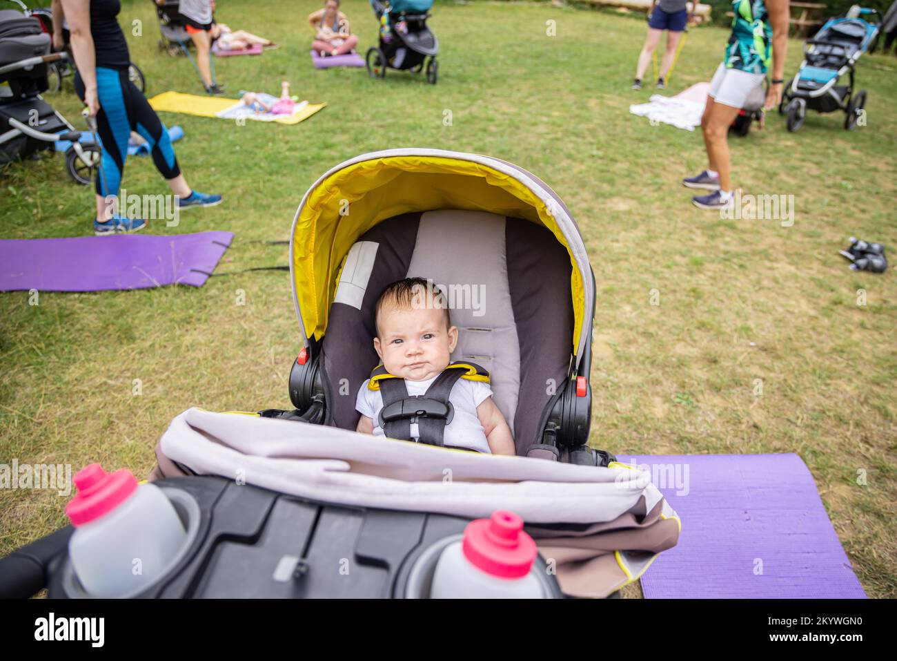 Ripresa grandangolare di un bambino che guarda dentro alla telecamera, mentre sullo sfondo altri genitori si stanno allenando in una sessione di gruppo sul campo d'erba Foto Stock