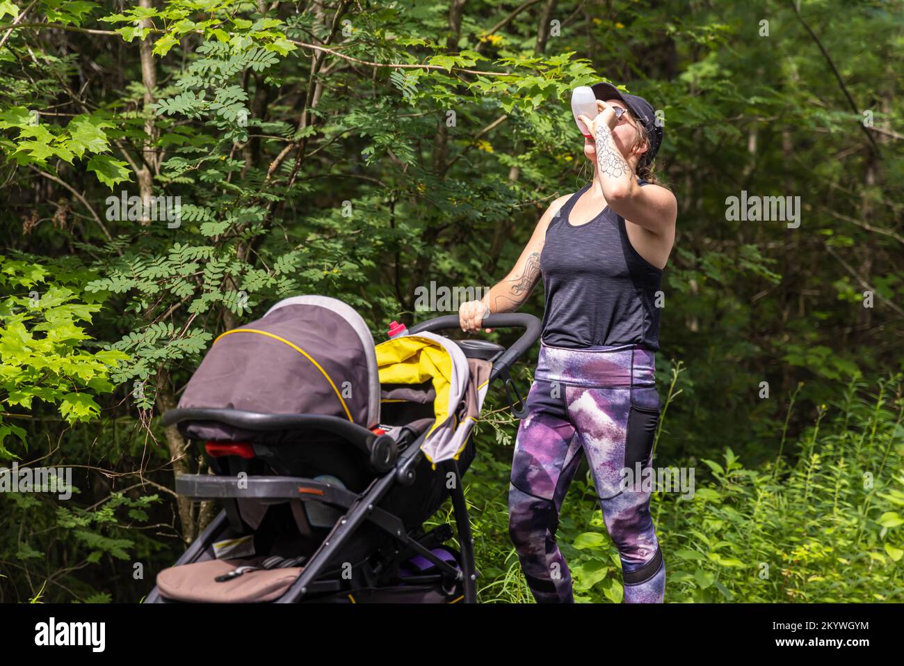 Foto ravvicinata della madre vestita di abbigliamento sportivo che beve dal biberon mentre cammina nell'ambiente naturale con il suo bambino all'interno del passeggino Foto Stock