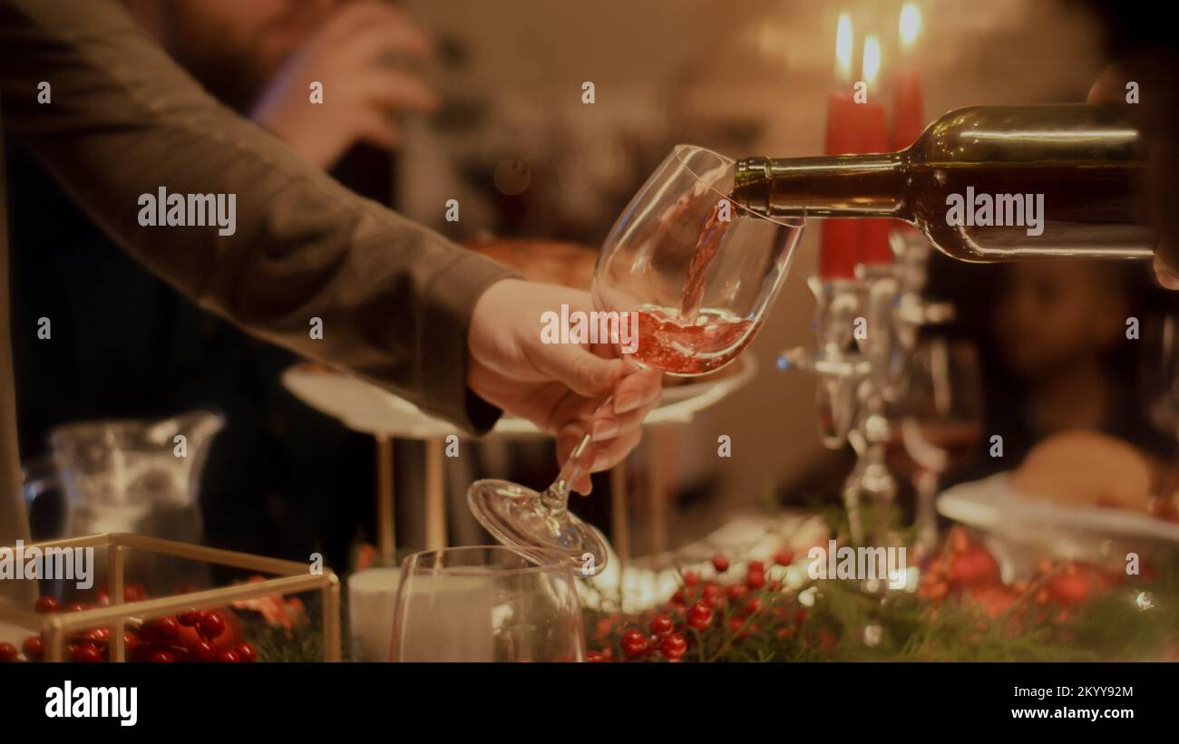 Un primo piano di un uomo versa il vino in un bicchiere di vino. La famiglia festeggia Natale, Capodanno o il giorno del Ringraziamento. Tavolo servito con piatti e candele. Calda atmosfera di famiglia cena di Natale a casa. Foto Stock