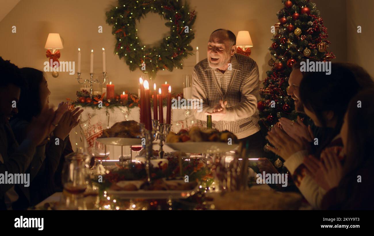 Il nonno anziano racconta scherzo o storia di vita alla sua grande famiglia multiculturale. Celebrano il Natale. Tavolo servito con piatti e candele. Calda atmosfera di famiglia cena di Natale a casa. Foto Stock