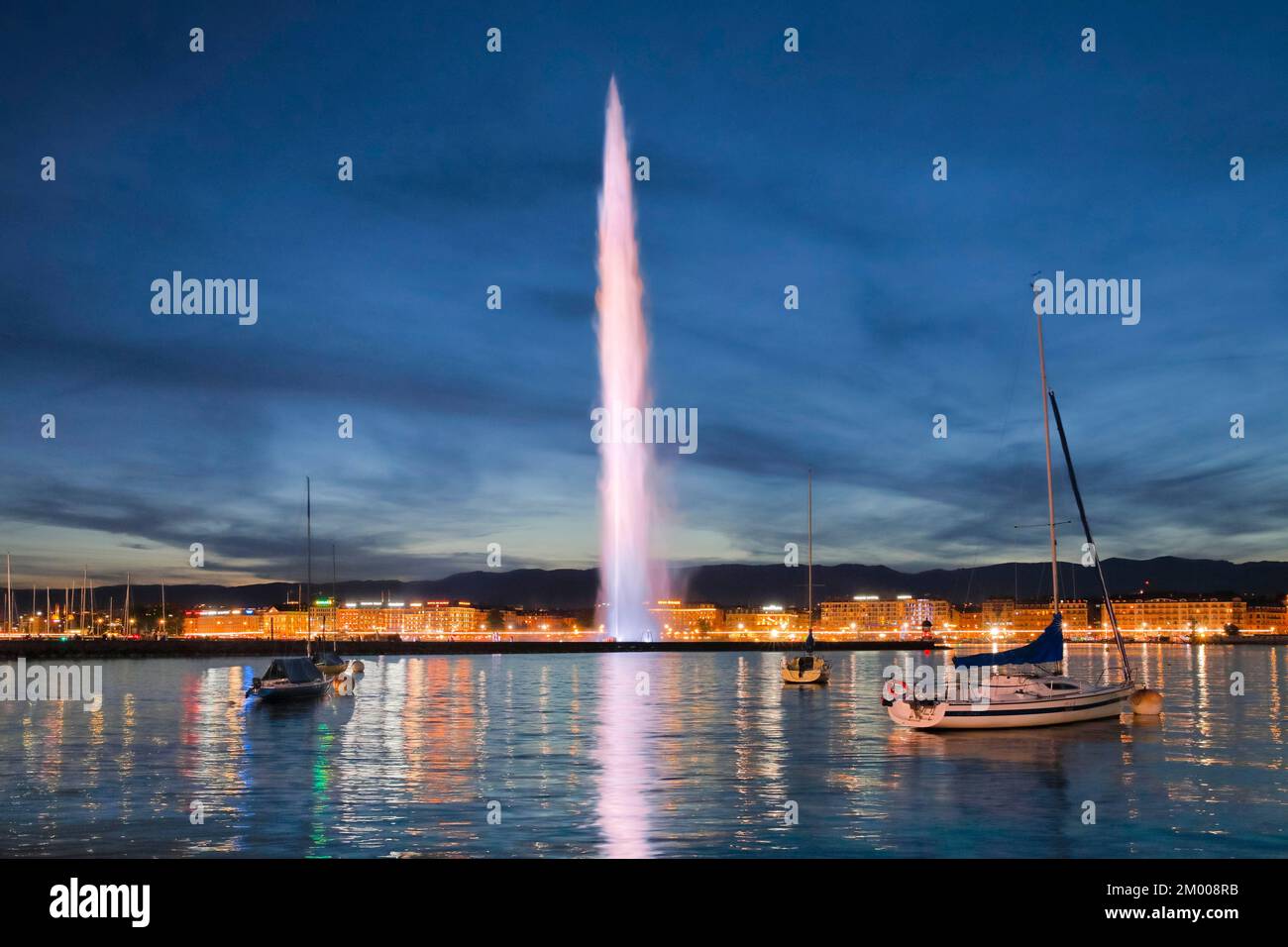 Il Jet d'eau illuminato di bianco al crepuscolo, punto di riferimento nel bacino del porto di Ginevra, con barche a vela in primo piano e la passeggiata sul lago illuminata di notte sul retro Foto Stock