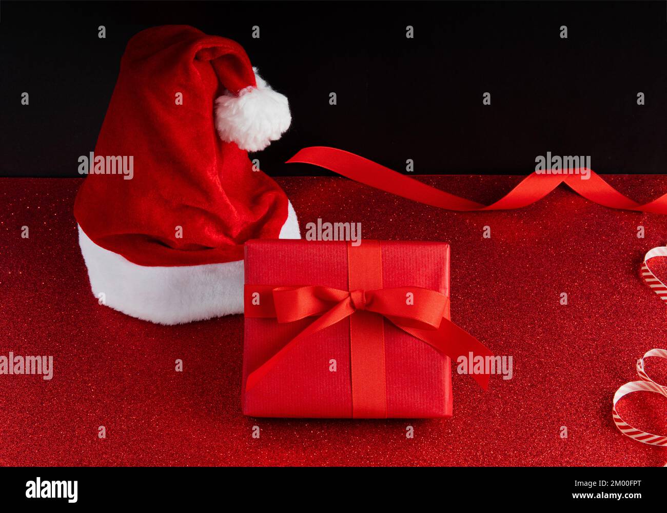 Sfondo di Natale con un cappello di babbo natale e una scatola regalo rossa con nastro su una superficie di glitter e uno sfondo scuro. Foto Stock