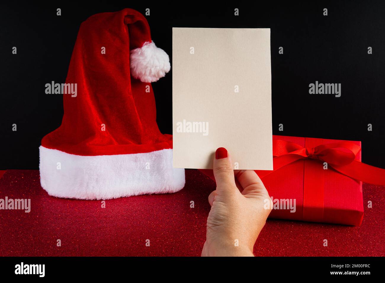 Mano della donna con le unghie rosse che tengono una cartolina vuota sopra un insieme di natale con un cappello di babbo natale ed una scatola rossa completa del regalo con il nastro Foto Stock
