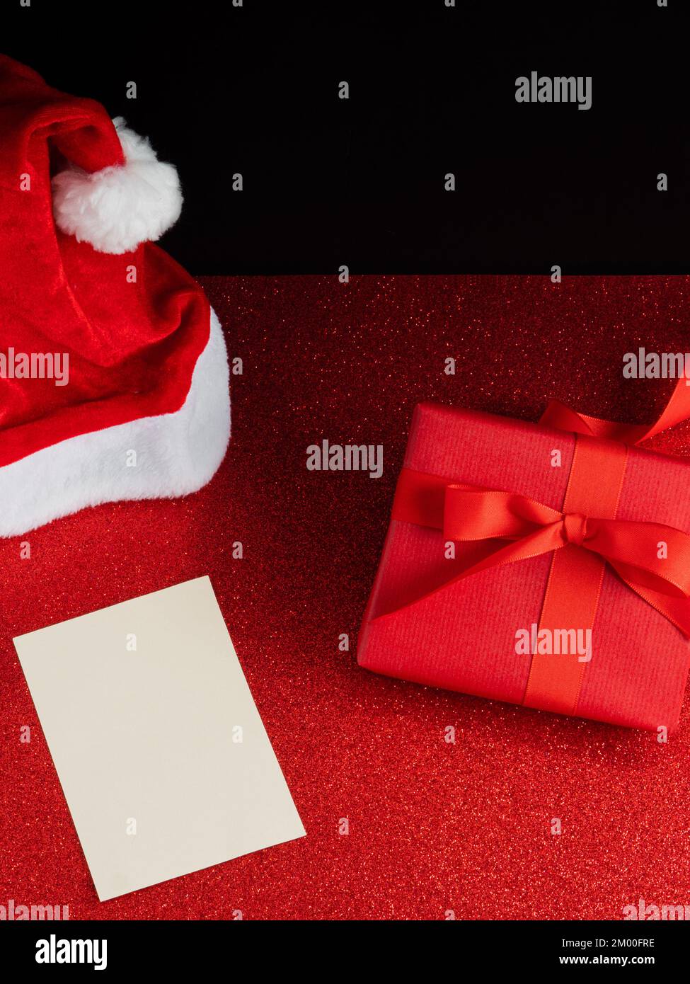 Cappello di Babbo Natale con un regalo rosso di lusso e una cartolina bianca per il mockup sullo sfondo di glitter Foto Stock