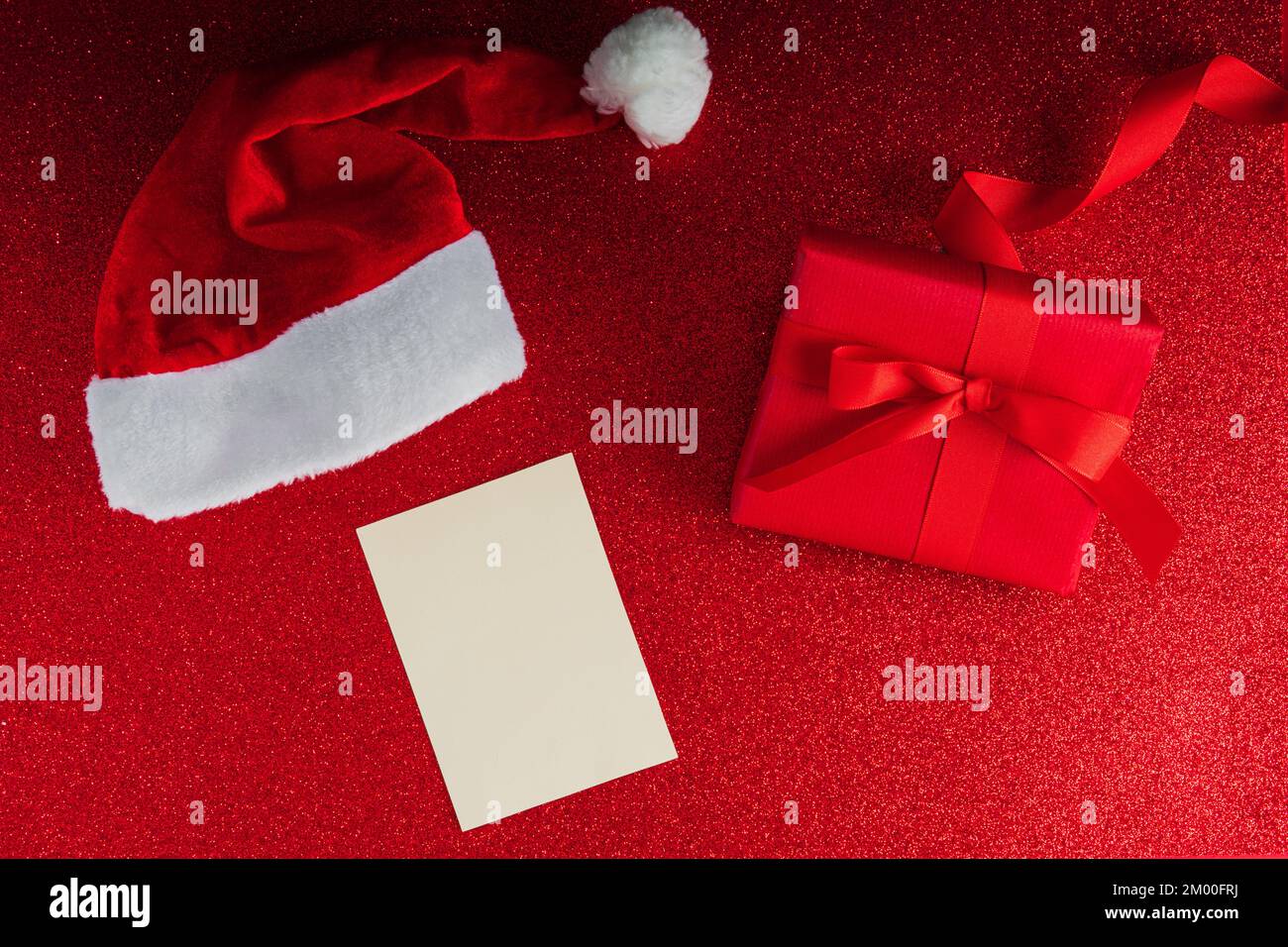 Cappello di Babbo Natale con un regalo rosso di lusso e una cartolina bianca per il mockup sullo sfondo di glitter Foto Stock