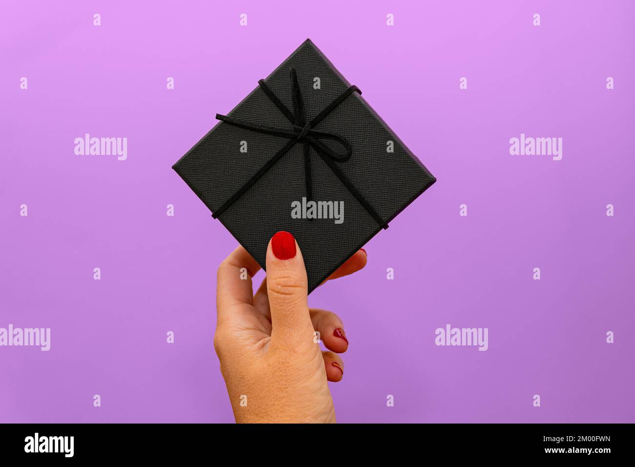 mano della donna con le unghie rosse che tengono il regalo nero sopra la parete viola Foto Stock