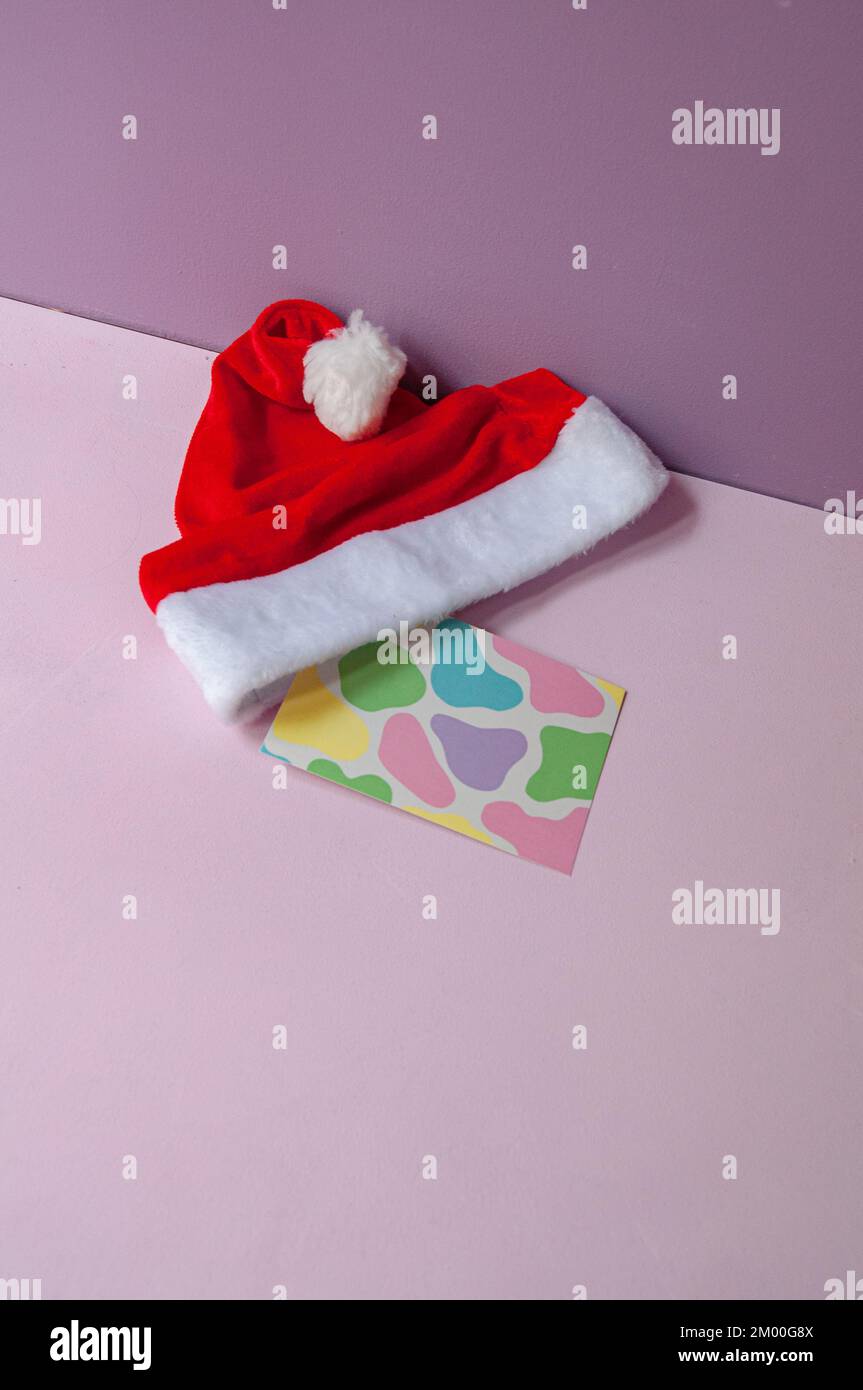 Cappello babbo natale con cartolina con disegno a mucca in colori pastello su sfondo lilla, in formato verticale con luce naturale Foto Stock