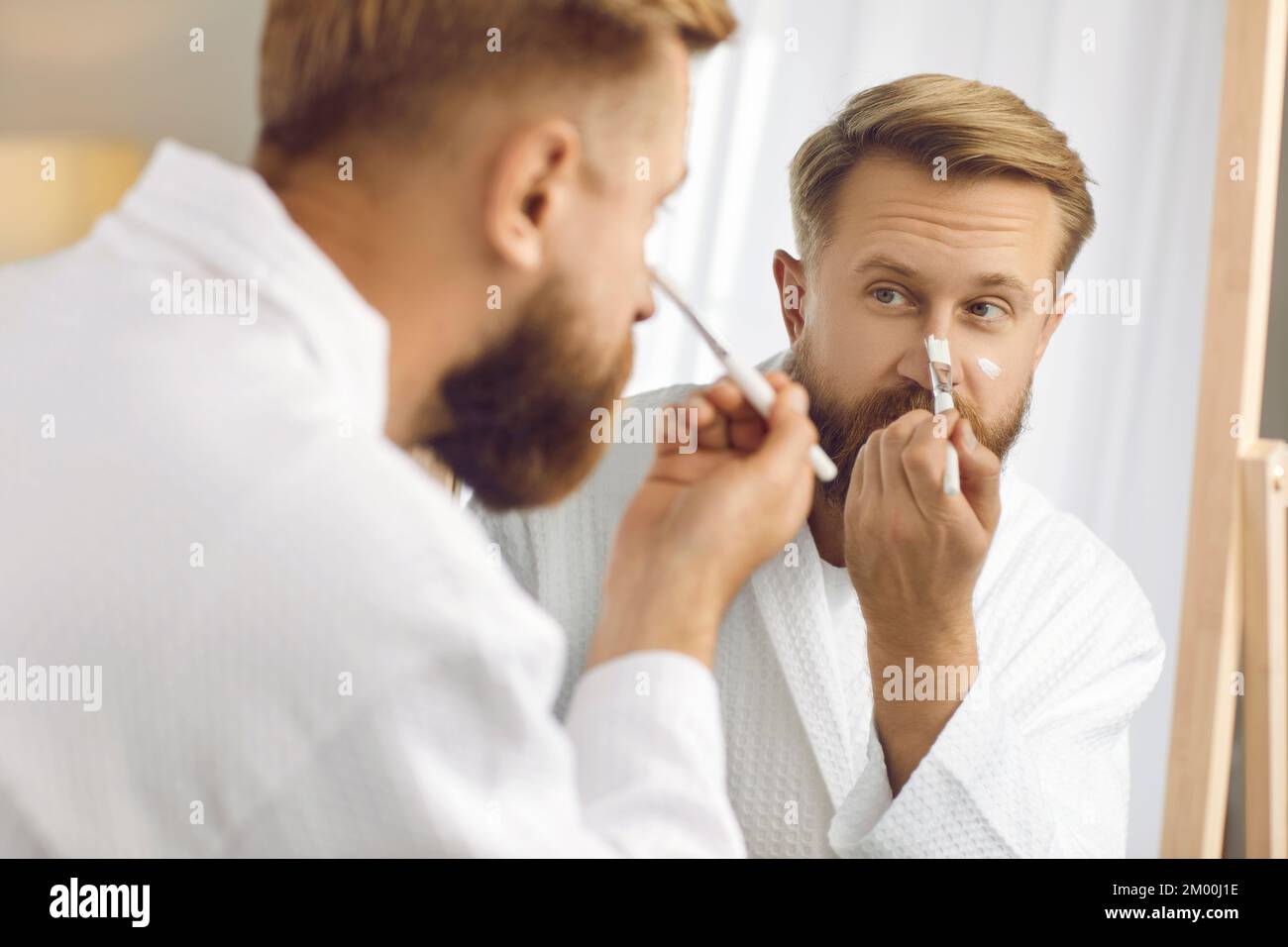 Bell'uomo bearded con pennello applica maschera idratante sul suo viso durante i trattamenti di bellezza a casa. Foto Stock