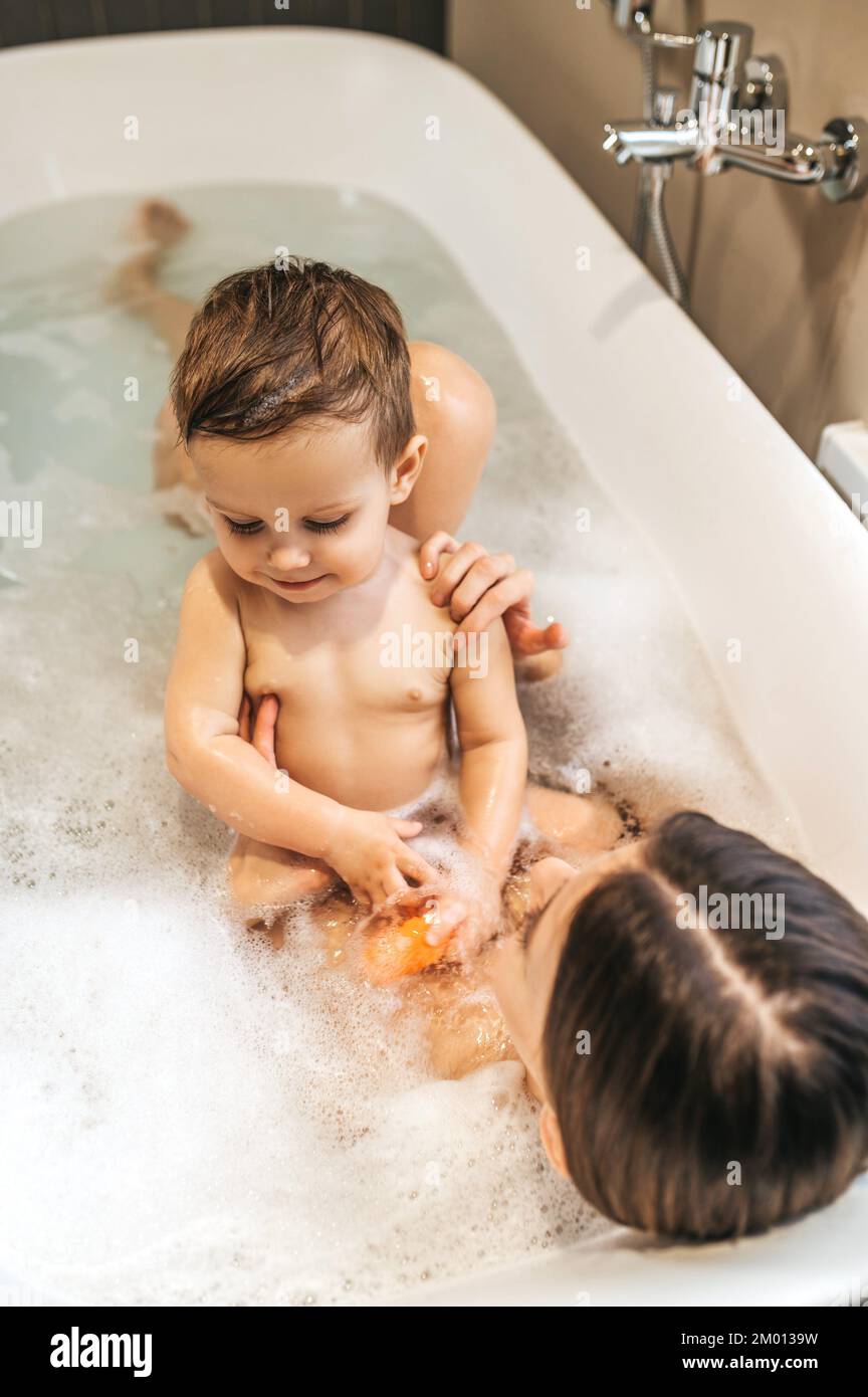 Preoccupandosi del genitore caucasico sdraiato nella vasca da bagno e supportante il suo bambino pacifico carino. Foto Stock