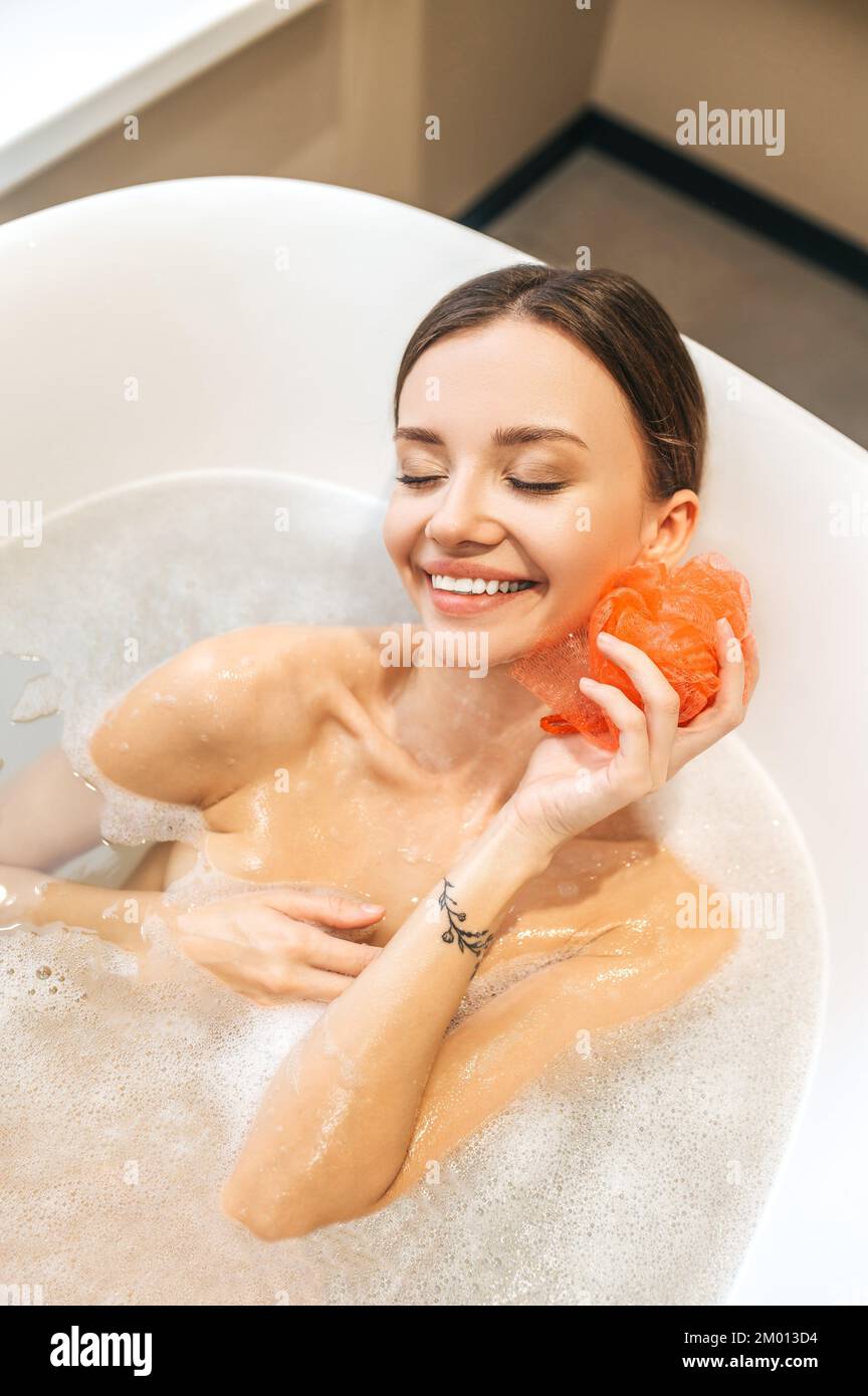 Bella donna felice sdraiata nel bagno e lavarsi con una loofah di nylon saponata. Foto Stock